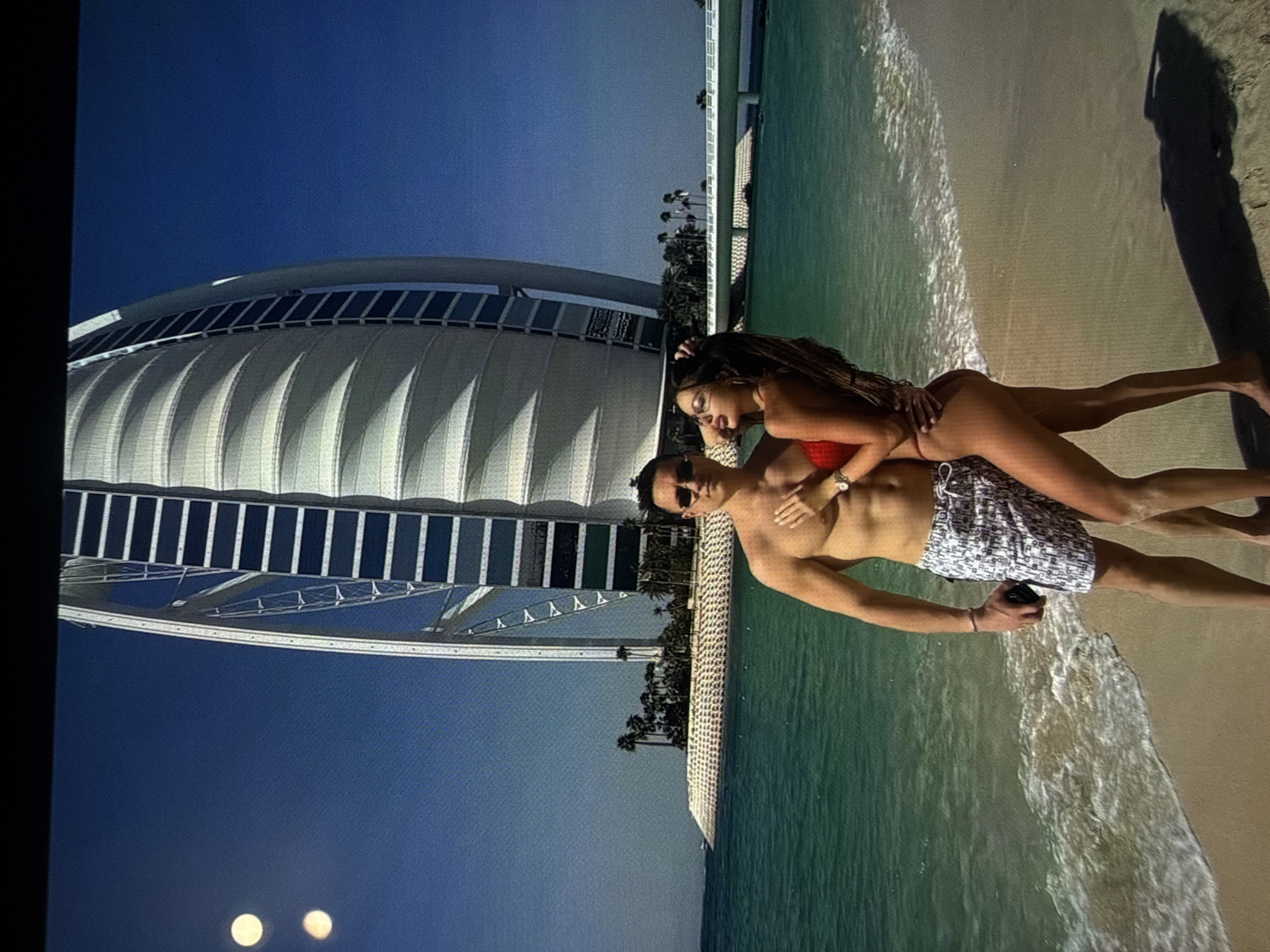 The image displays a man and a woman embracing on a sandy beach. The prominent background features the distinctive sail-shaped architecture of the Burj Al Arab hotel, located on an artificial island within turquoise waters, identifying the setting as Dubai, United Arab Emirates, not Varna, Bulgaria. A white bridge connects the artificial island. The man, shirtless and wearing patterned swim shorts and sunglasses, holds a dark object, likely a phone, in his left hand. The woman, wearing a red two-piece bikini and sunglasses, has her right hand placed on the man's chest and her left hand raised to her head; she is looking to her right with her mouth slightly open. Both individuals cast long shadows on the sand. Two bright, circular light artifacts are visible in the upper-left corner of the image.