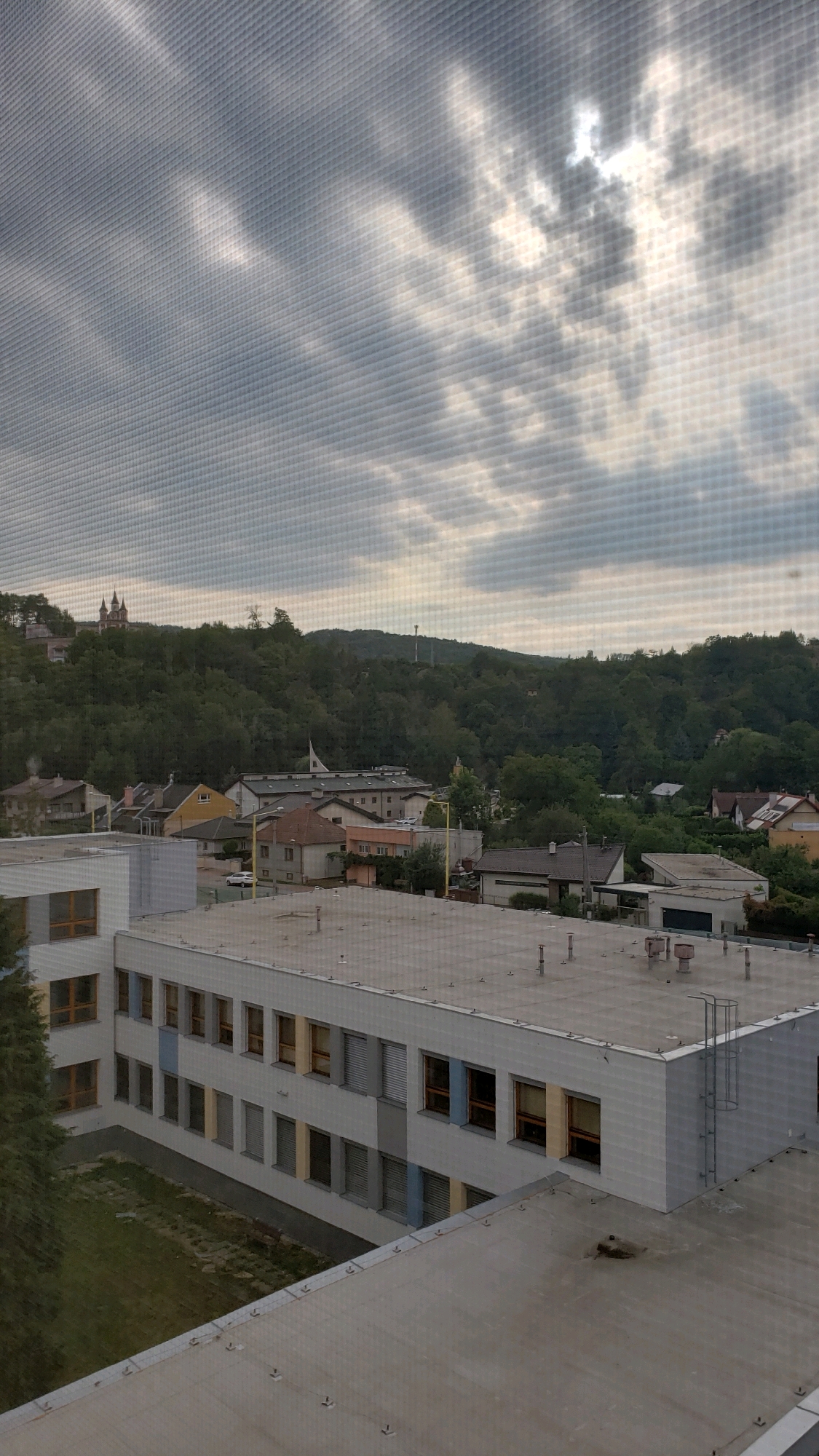 The image shows a view of buildings and a wooded hill through a screen. In the foreground, the flat roof of a building is visible, with some vents and other rooftop equipment. Partially in view is the side of another building, appearing to be a school or institutional building, with multiple windows. Beyond this building, a collection of houses and other structures forms a village or town. The background is dominated by a densely forested hill, upon which a church or religious building with twin spires can be seen. The sky above is overcast with dramatic clouds, and the sun is partially visible, breaking through the clouds. The overall impression is of a view from an elevated position overlooking a town nestled against a natural landscape. Based on the visible architecture, particularly the distinctive twin-spired church on the hill, this scene is likely located in Prešov, Slovakia.