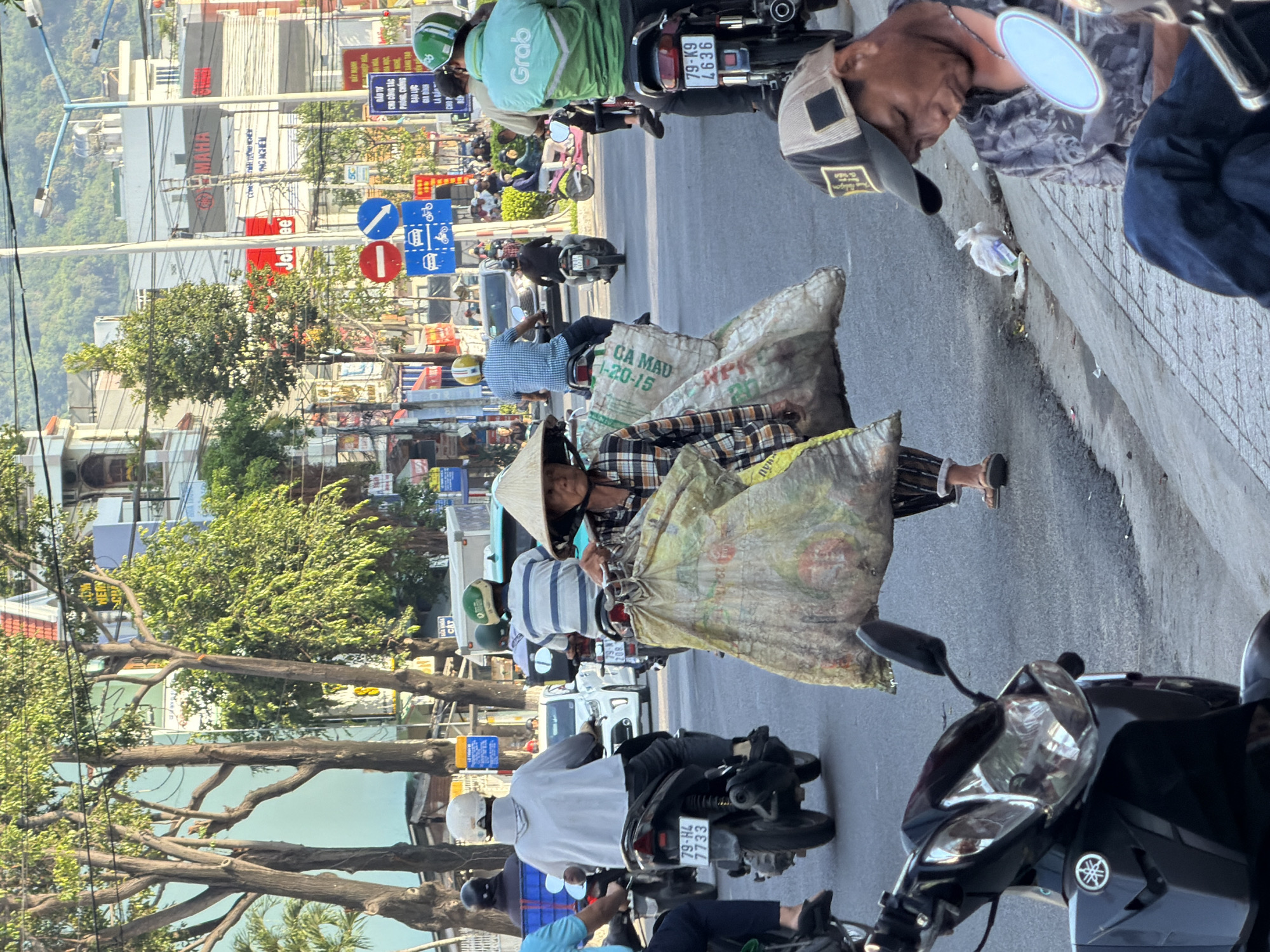 This image depicts a bustling street scene in Bac Nha Trang Ward, Vietnam, likely during daytime, given the strong sunlight and shadows. The foreground features several motorcycles, one of which is black and parked, its headlight prominently visible with a Yamaha logo. To the right, a man wearing a patterned shirt and a baseball cap is seated, looking down, with a motorcycle mirror reflecting light nearby.
In the mid-ground, a woman wearing a traditional conical hat and a plaid shirt is walking, carrying two large, bulging sacks that appear to be filled with goods. She is dressed in dark pants and sandals. Behind her, a busy street is filled with various vehicles, including motorcycles, scooters, and a white car. Several individuals are seen riding motorcycles, some wearing helmets. One person is wearing a green jacket with "Grab" logo, suggesting ride-sharing services are active.
The background shows a street with buildings, overhead wires, and traffic signs. Trees line the sides of the road, and distant hills are visible under a clear sky. The overall atmosphere is one of everyday urban life, with commercial activity and transportation clearly evident. The presence of multiple motorcycles and the general congestion indicate a typical busy street in a Vietnamese city.
