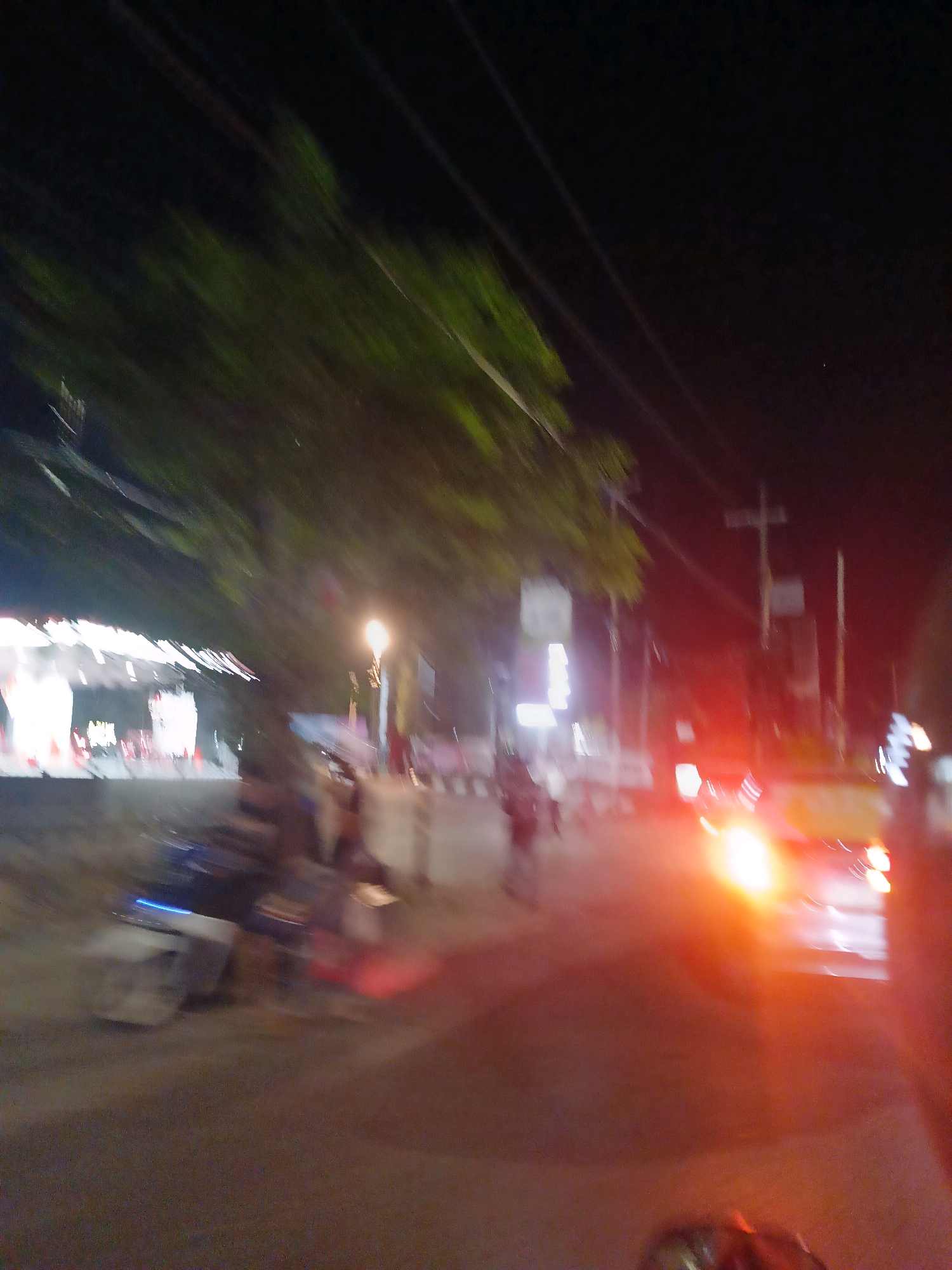 This is a night-time, motion-blurred photograph taken on a street in Makassar, Indonesia. The scene captures elements of a typical urban street at night, with artificial lighting from buildings and streetlights.
On the left side of the image, there appears to be a building or structure with bright, illuminated signage and what looks like a covered walkway or awning. Several objects that resemble traffic cones are visible near the edge of this structure. Further to the left, there are blurred shapes that could be people or vehicles.
The center and right side of the image show a street with traffic. A prominent feature is a bright, yellowish light source, likely the headlights or taillights of a vehicle, which is heavily blurred due to motion. Several other blurred lights, possibly from other vehicles and streetlights, are scattered across the scene, creating streaks of illumination against the dark background. There are also vertical elements that could be utility poles or streetlights.
The overall impression is one of a busy street scene at night, with the blur indicating movement, possibly from the photographer's perspective in a moving vehicle or from other traffic. The presence of illuminated buildings, streetlights, and vehicles strongly suggests an urban environment, consistent with a city like Makassar.