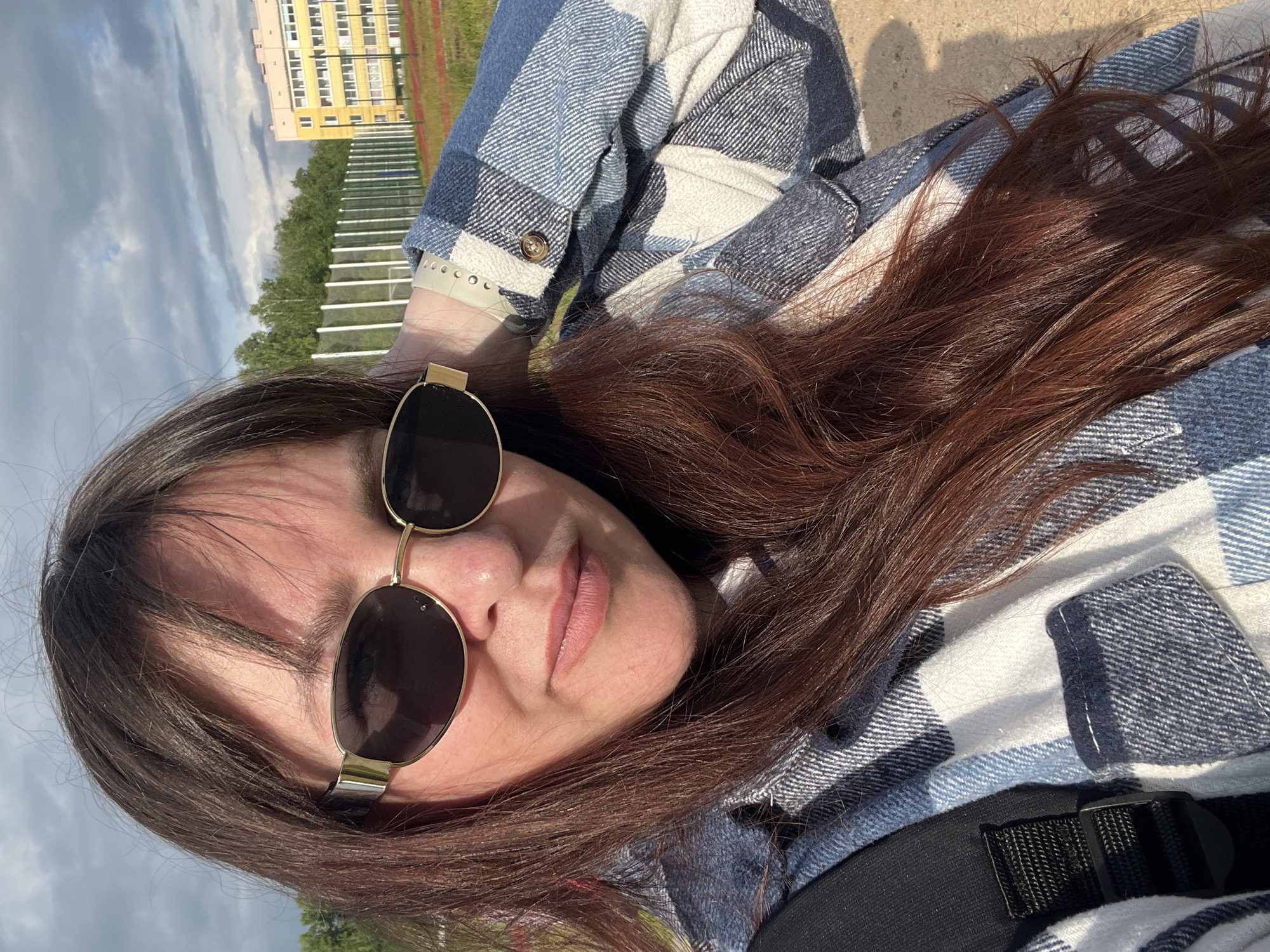 A woman is lying down outside, wearing sunglasses and a blue and white plaid shirt. Her head is tilted to the right, and her long brown hair is spread around her. The background features a building, trees, a fence, and the sky, suggesting an outdoor location. The scene appears to be in Noviy, Russia, based on location information.