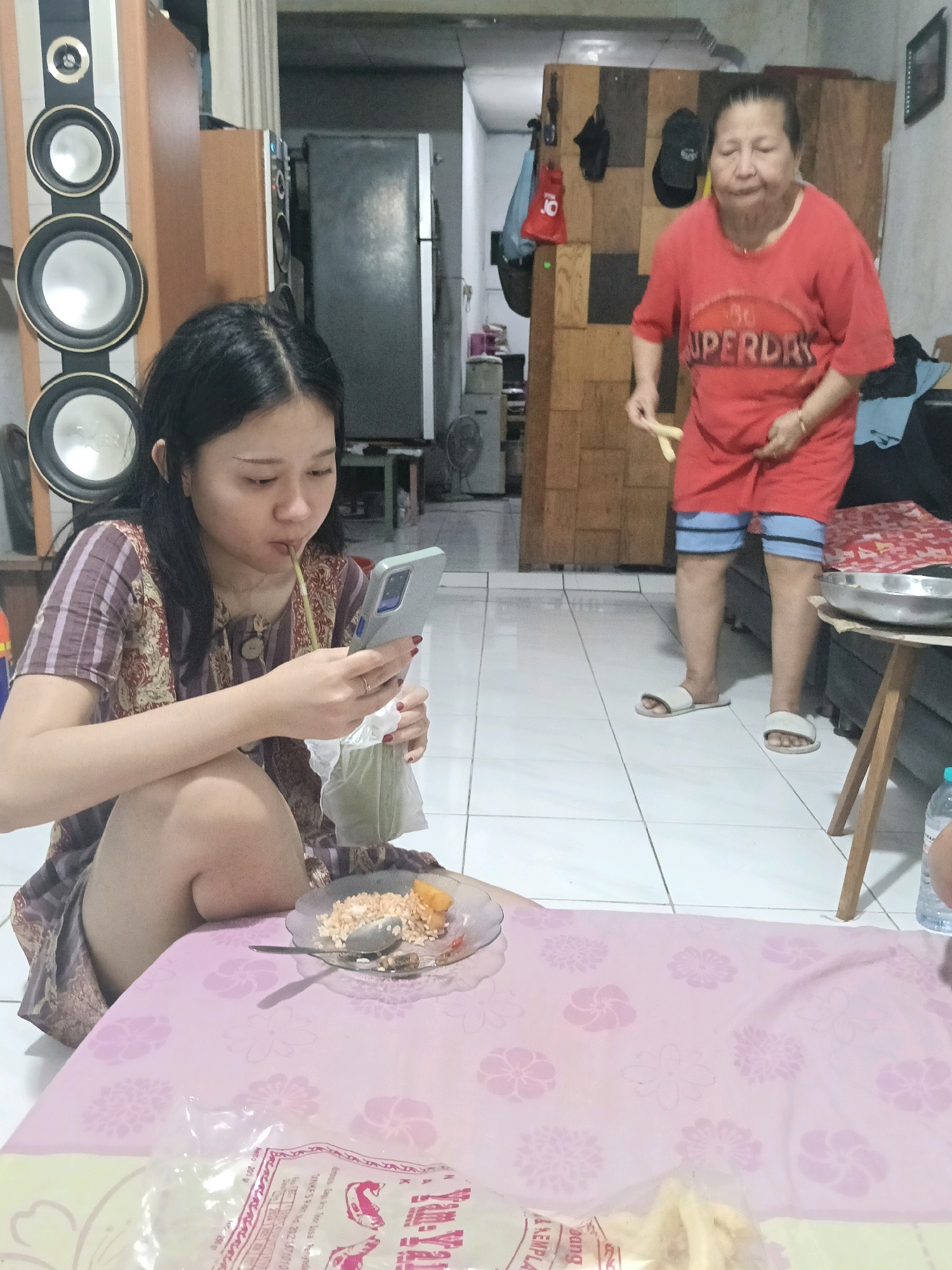 In a residential interior located in Jatimulya, Indonesia, a young woman with dark hair is seated on the floor, leaning to her left. She is wearing a purple and white striped dress and appears to be drinking from a bag with a straw while looking at her phone. In front of her, on a pink tablecloth with a floral pattern, is a plate with food, a spoon, and a plastic bag. To her right, a stack of large speakers is visible. Further back in the room, an older woman in a red t-shirt and blue shorts is standing and holding a yellow object. The room has tiled flooring and wooden cabinetry.