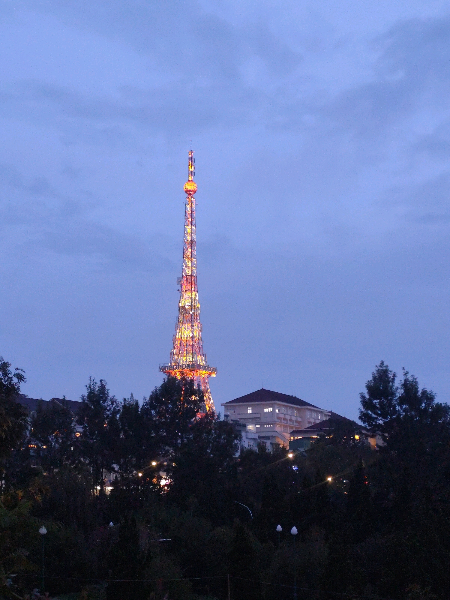 A telecommunications tower, illuminated by colorful lights, dominates the center of the frame, rising above a cluster of buildings and trees. The tower has a distinctive spherical section near its top. The sky above is a dusky blue, with some clouds present. The foreground is filled with the dark silhouettes of trees, with some artificial lights visible among them, suggesting a nighttime or twilight setting. Buildings with illuminated windows are visible behind the trees to the left of the tower. The scene is likely set in Da Lat, Vietnam, as the tower bears resemblance to the Da Lat Television Tower, a prominent landmark in the city.