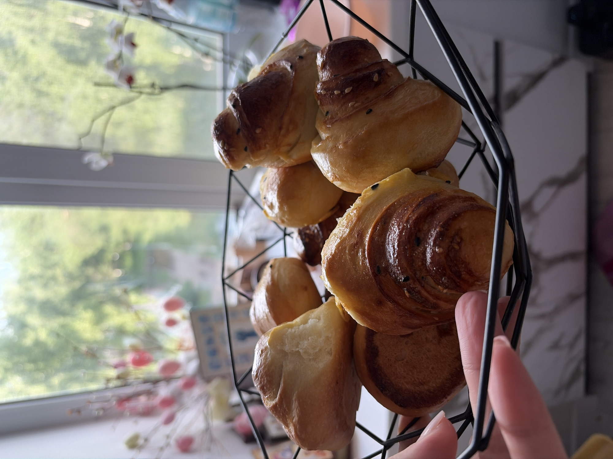 A person's hand is visible, holding a black wire basket filled with golden-brown baked goods, including croissants and round buns topped with sesame seeds. The basket is placed on a countertop. In the background, out of focus, is a window with a view of lush green trees and a hint of flowering branches, suggesting an indoor setting, possibly a home, near nature. The overall scene conveys a sense of domesticity and freshly baked treats. The location in Chaikovsky, Russia, is not discernible from the visual elements presented in the image.