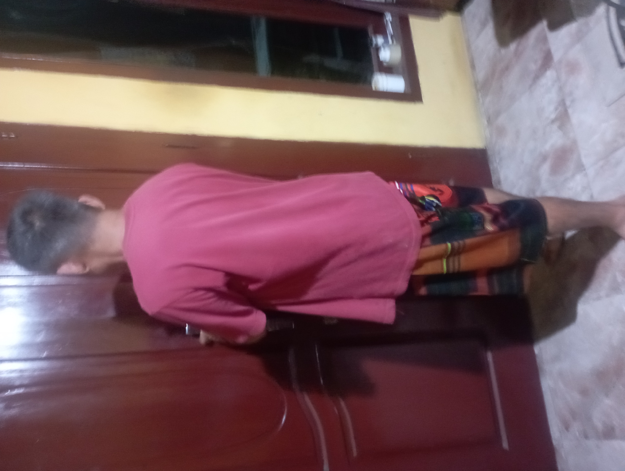 In the city of Maos Kidul, Indonesia, a person stands facing away from the camera, positioned between a reddish-brown door on the left and a yellow wall with a window on the right. The individual is wearing a pink t-shirt and patterned shorts with red, black, and green geometric designs. Their hair is short and dark. The door appears to be made of wood with some vertical grooves. The window on the right shows some objects on the sill, including a white cylindrical container and a small jar. The floor is tiled and has a marble-like pattern with some reddish-brown stains. The lighting in the scene is dim, casting shadows on the floor and wall. The overall environment suggests an indoor or semi-enclosed space, possibly a home or a similar building.