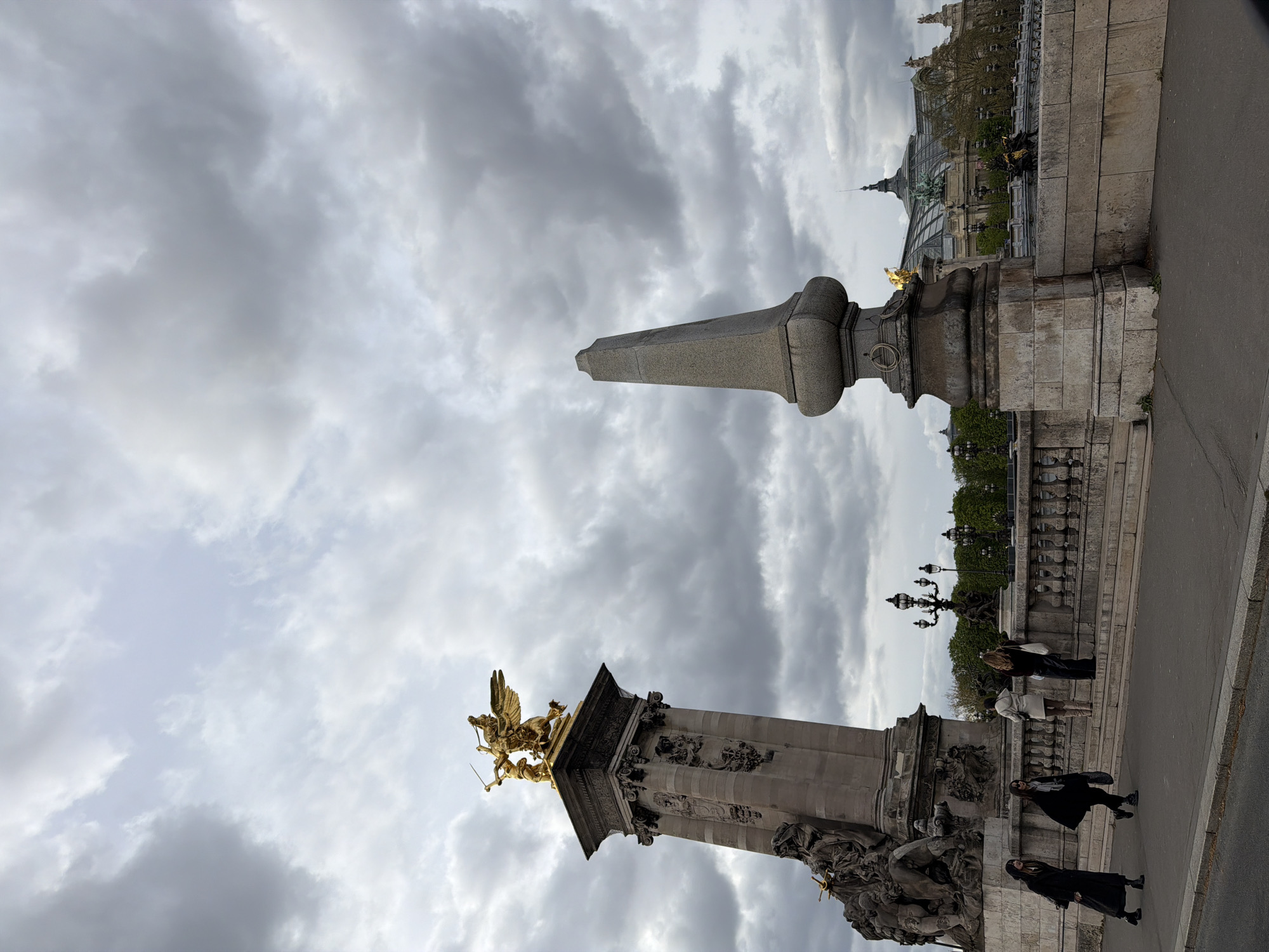 This is a view of the Pont Alexandre III in Paris, France. The scene captures the bridge's ornate stone pillars, one topped with a gilded sculpture of a winged figure on horseback. People can be seen walking along the bridge. The background includes the architecture of the Grand Palais, visible in the distance. The sky is overcast with grey clouds.
