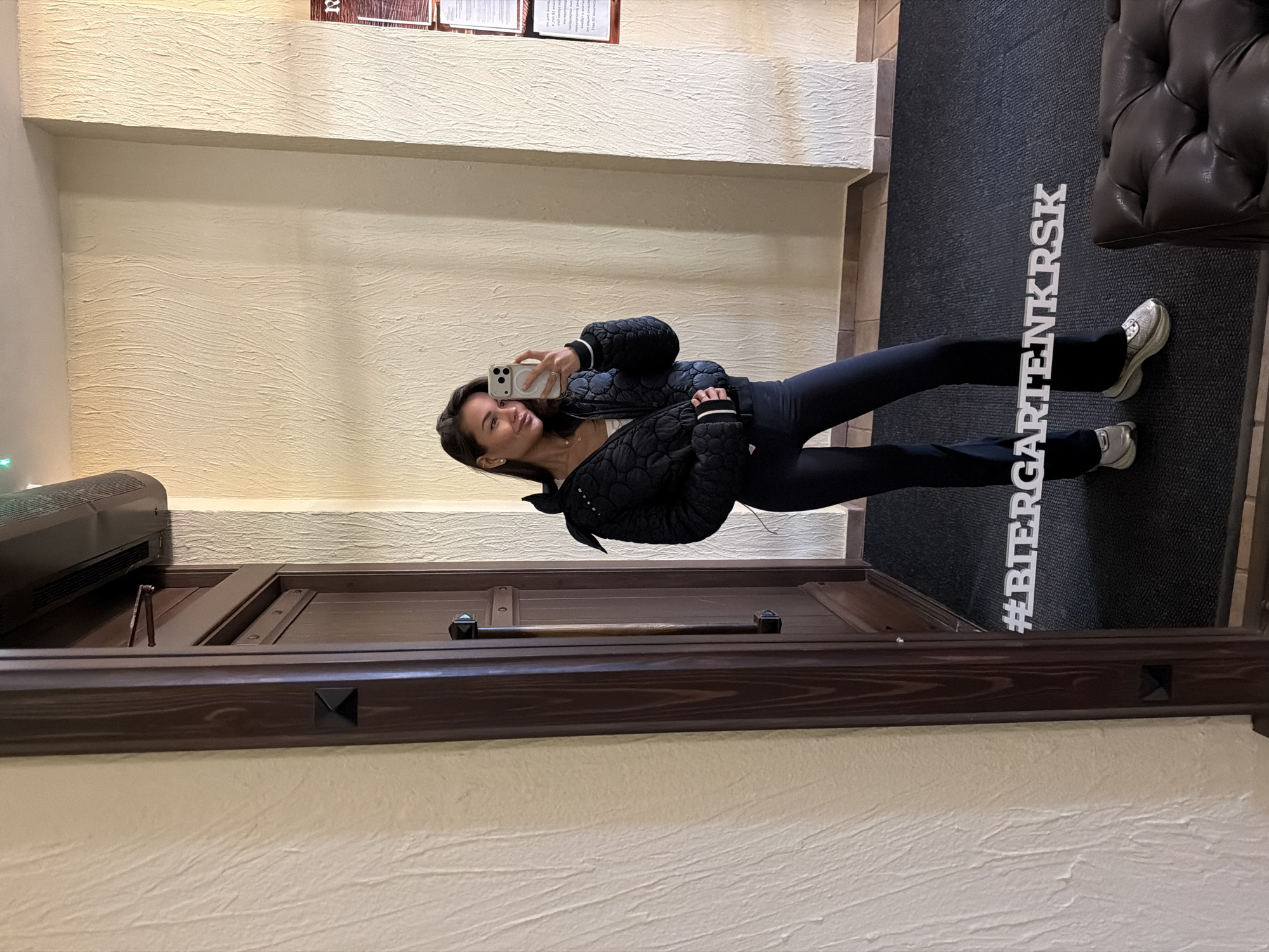 A woman stands in front of a full-length mirror, taking a selfie. She wears black leggings, a black quilted jacket, and light-colored sneakers. The mirror reflects a hallway with textured beige walls and a dark-colored entrance door. A black doormat with "#BIERGARTENKRSK" printed vertically in white is on the floor. A dark leather sofa sits to the right of the doormat. The location is Krasnoyarsk, Russia.