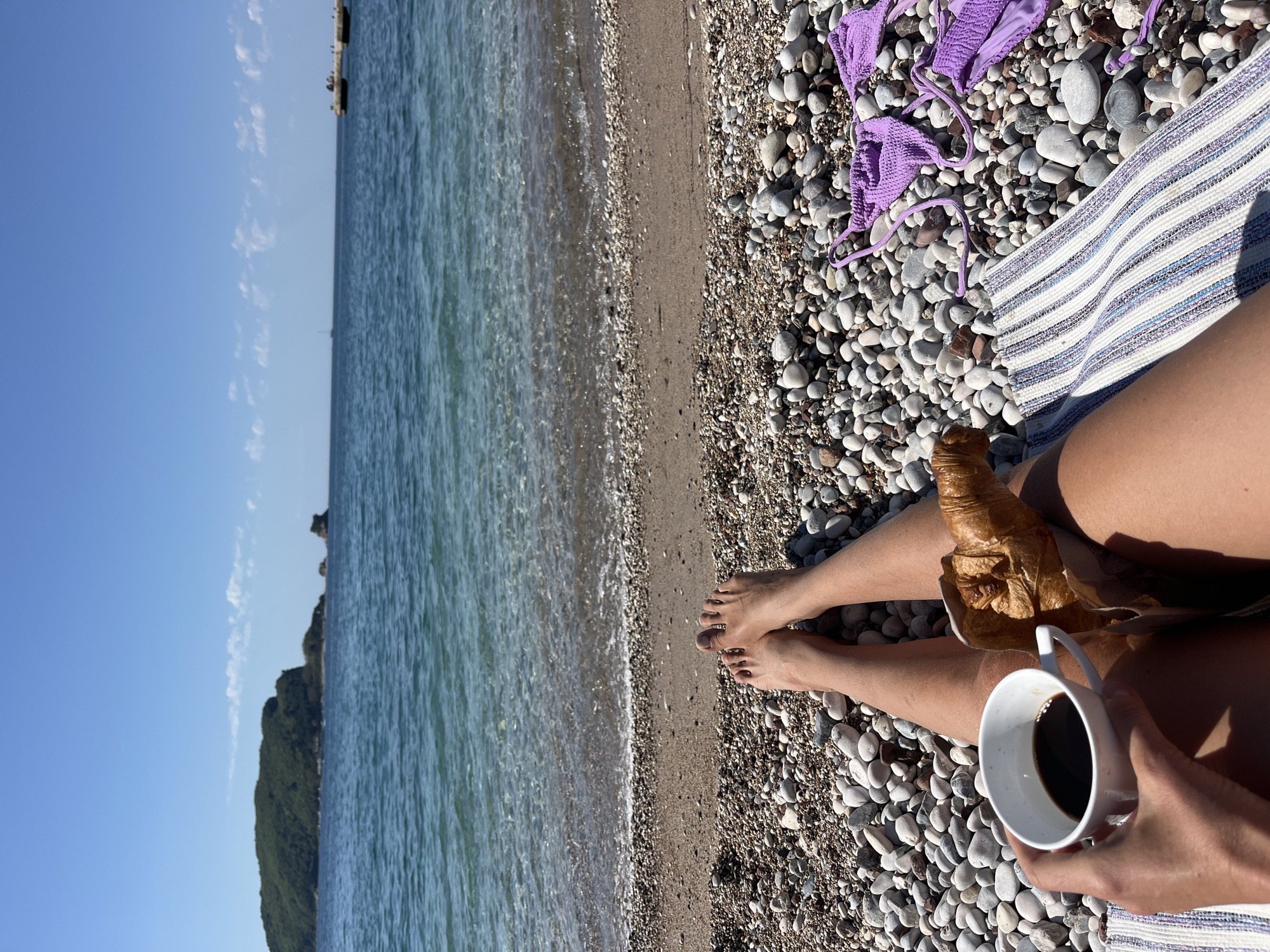 The image, taken from a first-person perspective looking down, depicts a serene moment on a pebble beach in Budva, Montenegro. The viewer's bare legs are extended towards the sea, resting on a striped mat or towel (white with blue and purple stripes). One hand holds a white ceramic mug filled with black coffee, while a golden-brown croissant, partially wrapped in brown paper, is placed on the other leg. Dark nail polish adorns the person's toenails.
To the right, a vibrant purple bikini top is spread out on the pebbled shore. The beach itself is composed of various sizes and shades of grey, white, and brown pebbles, with a narrow strip of darker sand or finer gravel closer to the water's edge.
Beyond the beach, the clear, blue-green sea extends to the horizon, with gentle waves lapping the shore. The distant background features a clear blue sky with a few scattered wispy clouds. On the left side, a dark green, rocky headland or mountain covered in lush vegetation juts out into the water.
The activity taking place is a peaceful breakfast or snack by the sea, enjoying the morning sun. The bright sunlight and shadows indicate an early morning setting, ideal for a relaxing start to the day. The scene evokes a sense of tranquility and holiday enjoyment. No text is visible in the image.