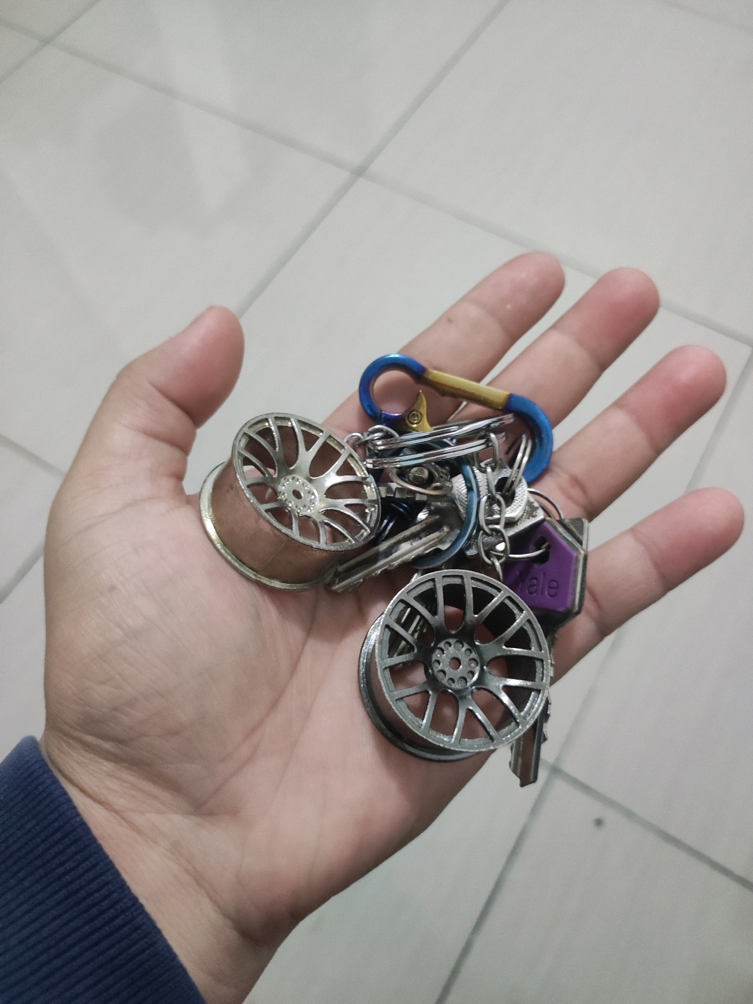 An overhead shot shows a person's left hand holding a cluster of keys. The keys are attached to a blue carabiner clip with a gold accent. Among the keys are at least three decorative keychain charms shaped like car wheels and one purple keychain fob. The hand is resting on a light-colored tiled surface, possibly a floor. The background is blurred and out of focus, showing more of the tiled surface. The location is not discernible from the image.