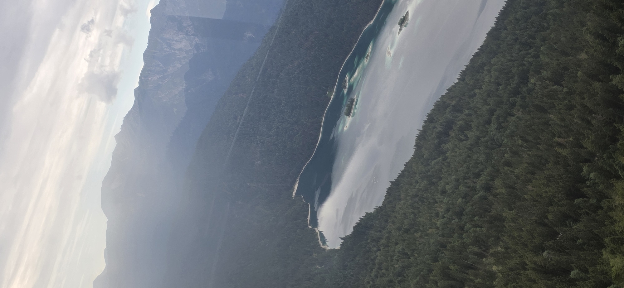 This is an aerial photograph of a pristine lake, likely in Grainau, Germany, surrounded by lush, dense evergreen forests. The water is a striking turquoise, with patches of lighter, sandy areas visible beneath the surface, suggesting shallow depths or underwater shoals. Several small, tree-covered islands dot the lake, adding to its picturesque beauty.
The landscape is dramatic, with steep, mist-shrouded mountains rising in the background. The mountains are characterized by rugged peaks and slopes partially obscured by atmospheric haze, giving the scene a sense of depth and scale. The lower slopes of the mountains, as well as the land bordering the lake, are covered in a dense canopy of dark green pine trees.
The sky above is a mix of soft, wispy clouds and clearer blue, hinting at a partly cloudy day. The light suggests it is either early morning or late afternoon, with a diffused glow illuminating the scene. There is no visible text in the image. The overall impression is one of natural beauty, tranquility, and remote wilderness. The perspective from above emphasizes the vastness of the forest and the clarity of the water.