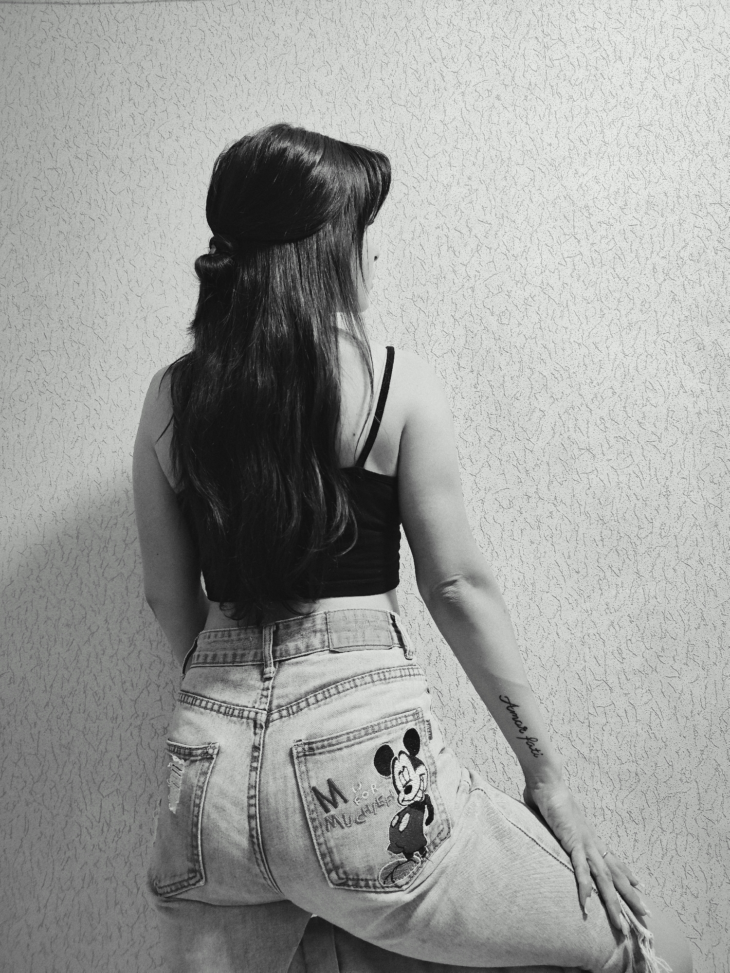 A black and white photograph shows a woman from the rear, positioned near a textured wall. She has long, dark hair, styled with a bun at the back of her head, and wears a dark tank top. Her light-colored denim jeans feature a Mickey Mouse design on the right back pocket. A tattoo is visible on her right forearm. Her right hand rests on her right leg. The location is not Фоки, Russia, as this information cannot be determined from the image.