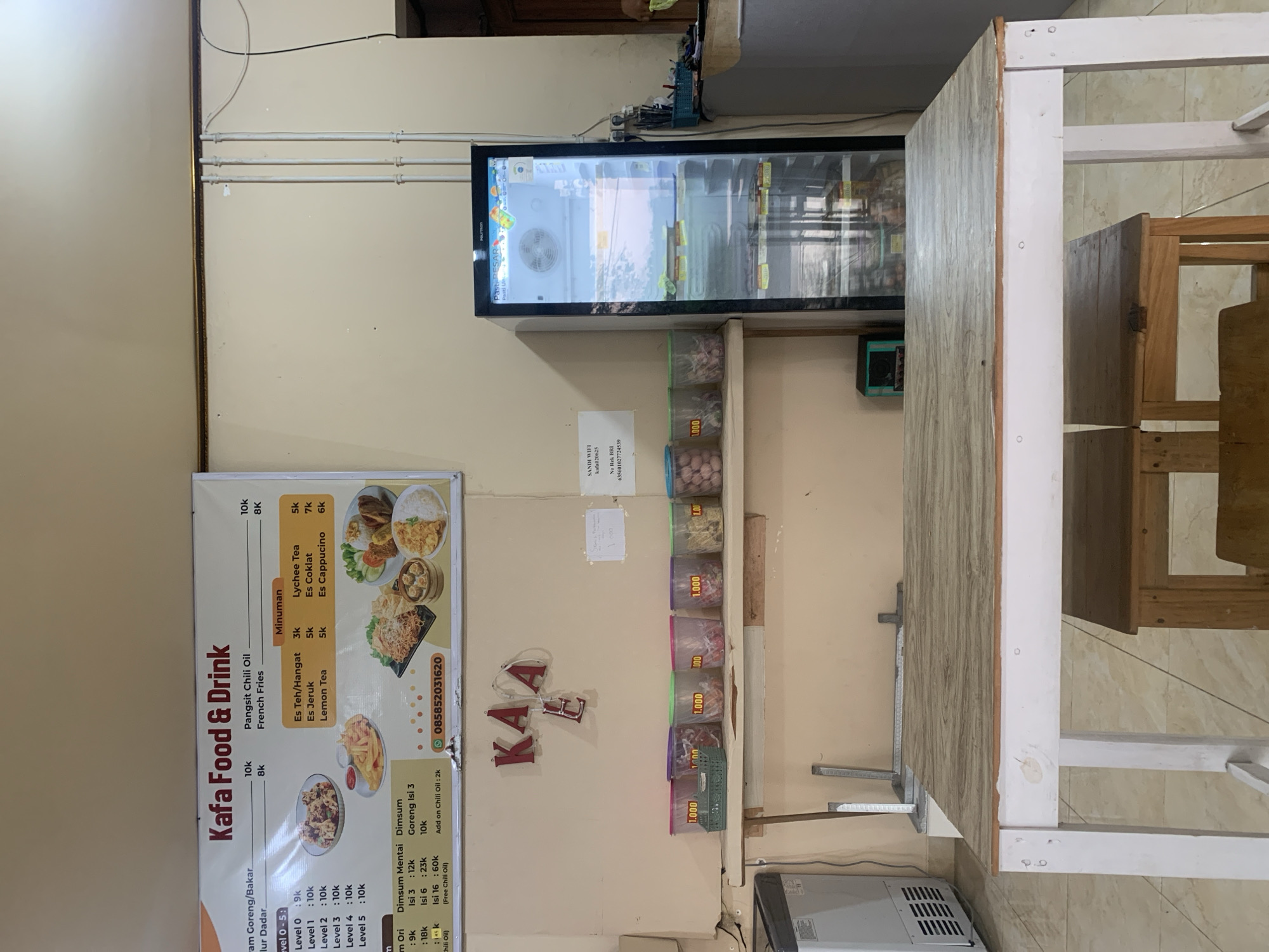 The image depicts the interior of a casual dining establishment, possibly a warung or small restaurant, in Jungke, Indonesia. A prominent menu board is displayed on the wall, listing food and drink items with prices. To the right, a glass-fronted refrigerator is filled with various food products. In the foreground, a simple wooden table and chairs are set up, suggesting a dining area. The walls are painted a neutral color, and the flooring appears to be tiled. Several plastic containers with labels indicating prices are placed on a shelf beneath the menu board. The overall ambiance suggests a functional and unpretentious eating space.