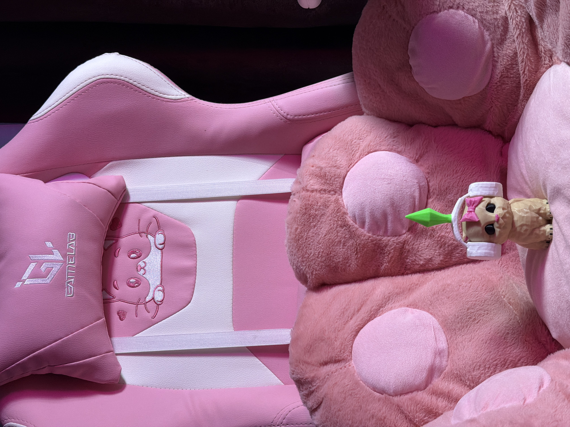 A close-up shot showcases a pink gaming chair with white accents and a pink lumbar pillow embroidered with a cat face and pink hearts. A small, tan figurine of a cat wearing white headphones with a pink bow and a green Sim-like plumbob on its head is positioned in the center of the chair's plush, pink armrests. The background is a dark curtain. The scene is indoors, in a room with no discernible urban features.