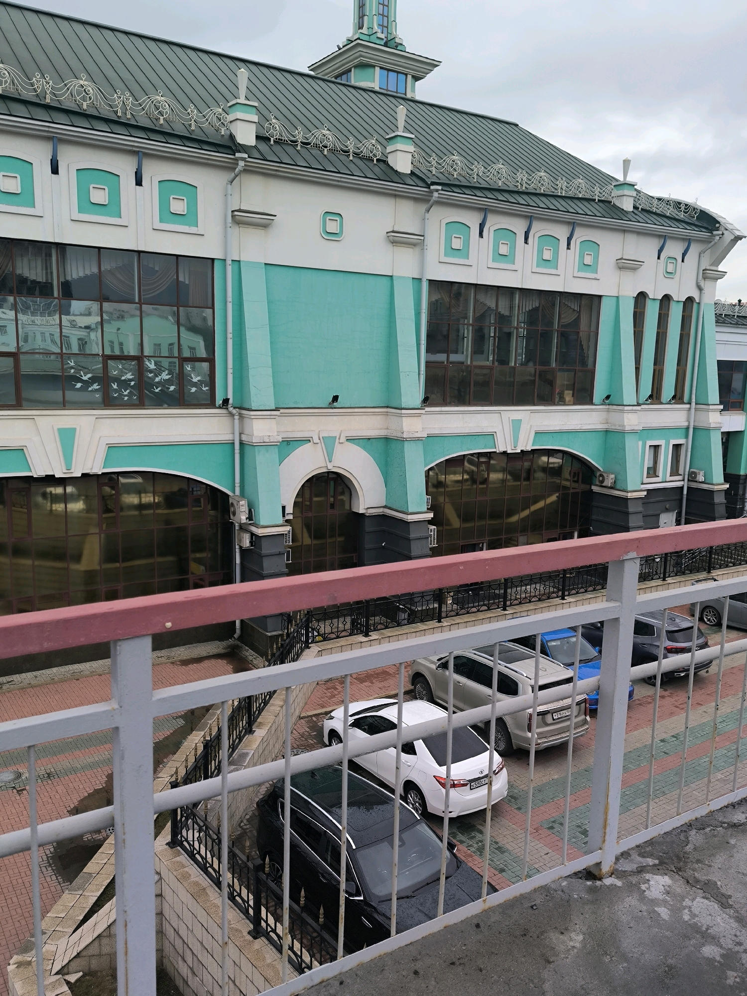 The image shows the Novosibirsk Central Railway Station, a prominent landmark in Novosibirsk, Russia. The building features a distinctive teal and white facade with large windows reflecting the cloudy sky. A metal fence runs along the foreground, partially obscuring a view of several parked cars below. Among the vehicles, a white sedan and a silver SUV are clearly visible, along with a blue car and a dark-colored SUV. The ground beneath the cars is paved with red and green bricks. The overall scene is urban and depicts a typical day at a busy transportation hub.