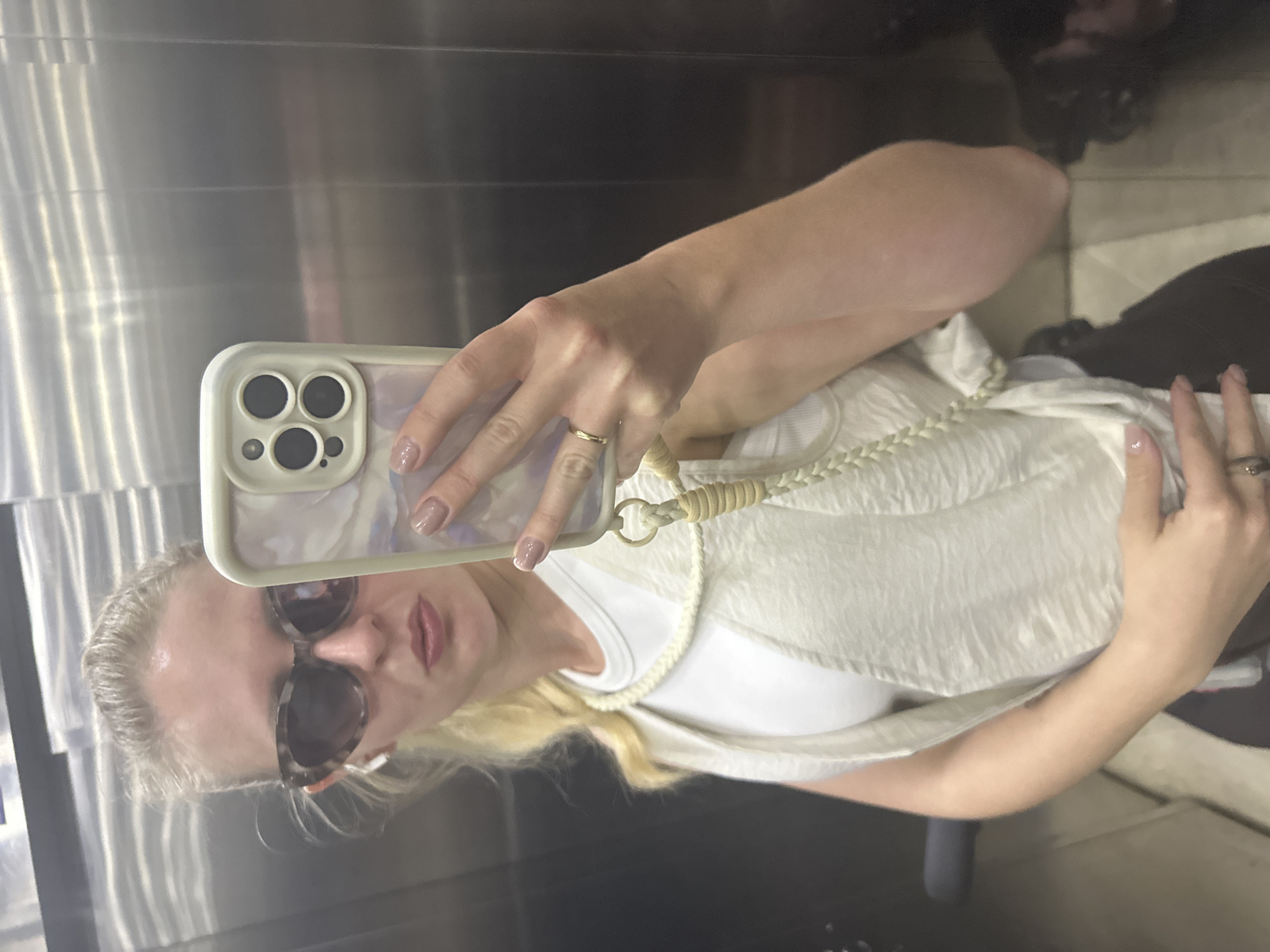 A selfie, rotated for ease of viewing, depicts a fair-skinned blonde woman in an elevator, likely in Petah Tikva, Israel (based on the background and metadata if available). She wears sunglasses and a white top. She is holding a smartphone with a triple-lens camera and a light-colored case, taking the picture. Her hands are visible, one holding the phone and the other resting on her lap. Both hands feature manicured nails. A gold ring is visible on her ring finger. The elevator has stainless steel walls. The floor is visible in the reflection. There is an obscured dark mass possibly a bag or the back of a seat in the lower right corner of the image.