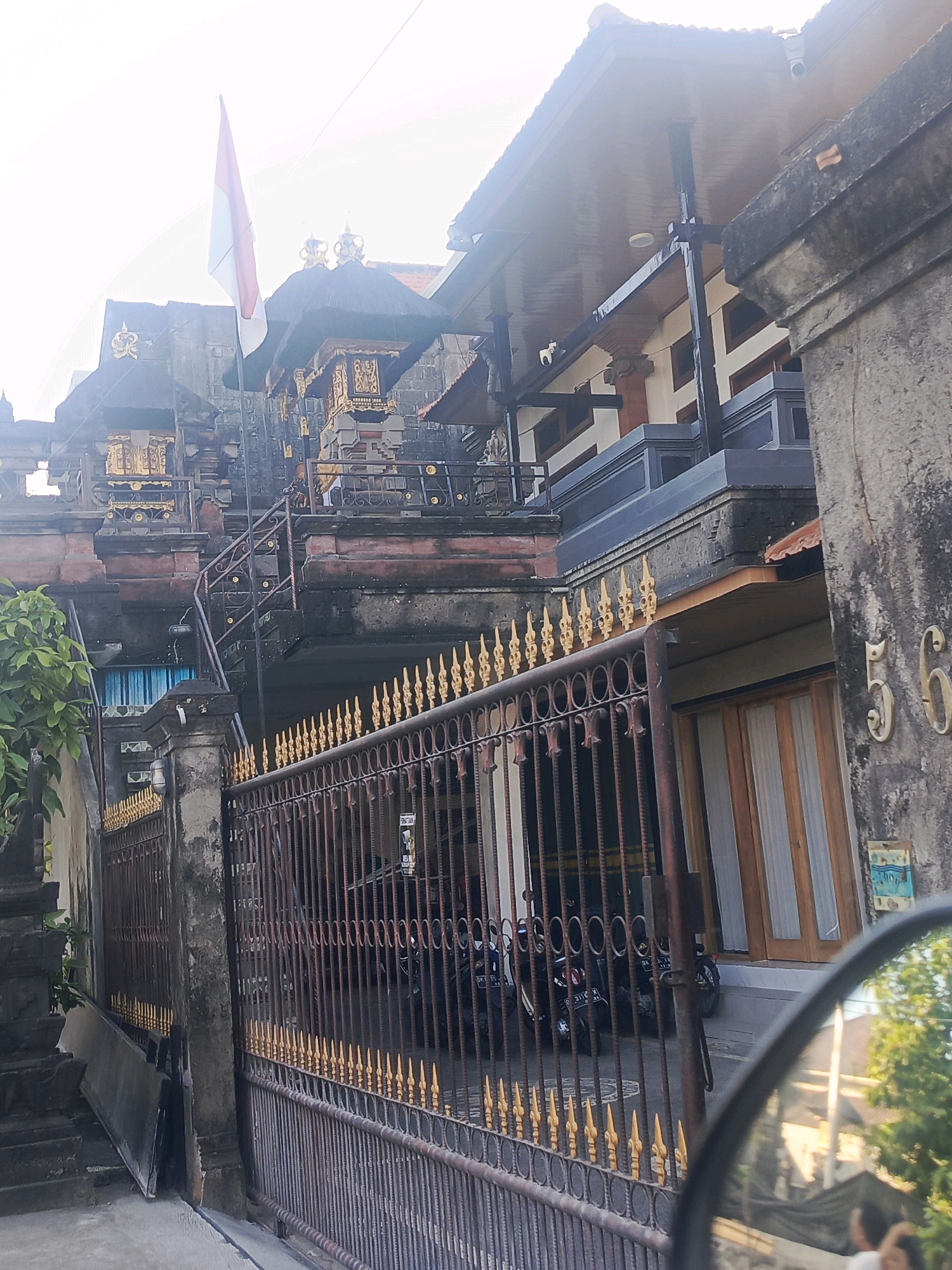 The image depicts a traditional Balinese building in North Kuta, Indonesia, characterized by its intricate architecture and ornate decorations. The building features multiple levels, with layered roofs and elaborate stone carvings. A red and white flag, likely the Indonesian flag, is prominently displayed on a flagpole.
In the foreground, a dark metal gate with golden decorative finials separates the street from the property. Behind the gate, several motorcycles are parked. To the right of the gate, a stone wall has the numbers "56" embossed on it. A wooden-framed door with vertical white blinds is visible on the right side of the building.
The environment appears to be a sunny day, with natural light illuminating the scene. There are no people clearly visible in the image, although a reflection in what appears to be a car's side mirror in the lower right corner shows a blurred image of a person's head and shoulder. The overall impression is one of a culturally rich and well-maintained residential or temple-like structure.