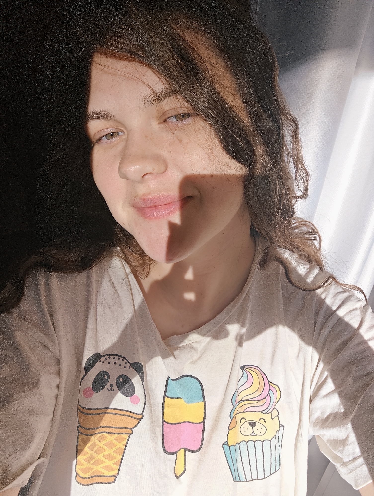 A young woman with long, wavy brown hair and light brown eyes smiles gently at the camera in a sunlit indoor setting. Her face is partially illuminated by bright natural light streaming from the right, casting distinct, sharp shadows across her features, notably down the center of her face and on her chin. A dark, shadowed area occupies the left side of the frame, contrasting with the well-lit right side where white or light-colored curtains are visible.
She is wearing a light-colored t-shirt adorned with three colorful cartoon designs. On the left, a panda character is depicted as an ice cream cone, complete with black ears, rosy cheeks, and small black dots above its eyes, set in a waffle cone. To its right, a striped popsicle features blue, yellow, and pink segments. Further right, a cheerful yellow pug with closed eyes and a slight smile is illustrated as a cupcake, topped with swirled, rainbow-colored frosting in shades of pink, yellow, blue, and purple, sitting in a light blue cupcake wrapper.
The strong, directional sunlight suggests the time of day is either early morning or late afternoon, creating a warm and relaxed atmosphere. Her expression is calm and pleasant. The scene depicts an everyday moment indoors, likely in a domestic environment in Chaikovsky, Russia. No discernible text is visible apart from the cartoon designs on her shirt.