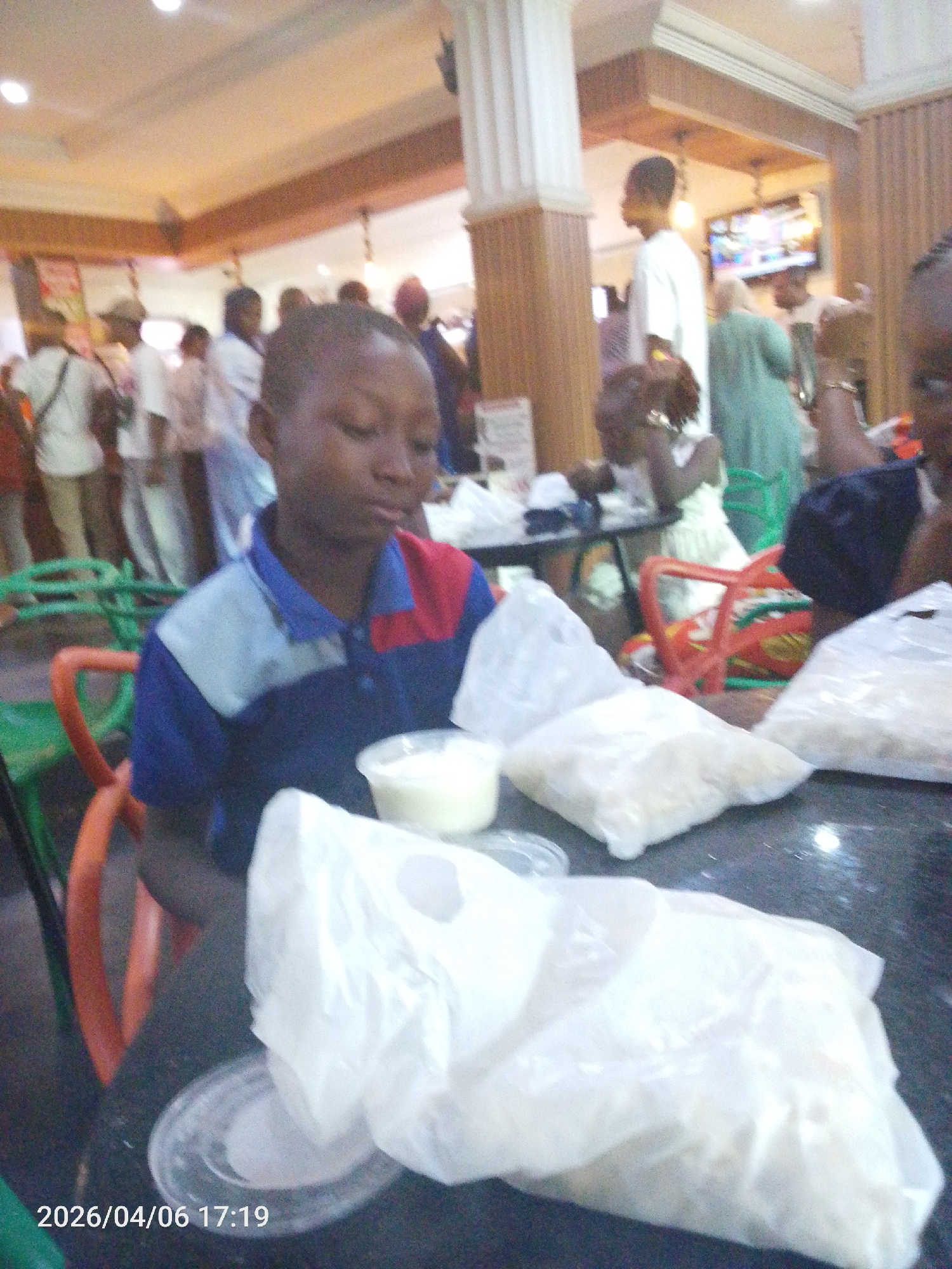 A medium shot captures a child seated at a table in what appears to be a food court or cafeteria-like setting. The child, of African descent, wears a blue and red collared shirt. Several translucent plastic bags, likely containing food items, are arranged on the table in front of the child. A small, clear plastic container holding a white substance is also visible on the table.
In the background, numerous individuals are seen milling about. Some are seated at tables, suggesting the area is a communal dining space. A white pillar adds to the interior architecture.
The timestamp "2026/04/06 17:19" is displayed at the bottom left corner of the image. While the prompt specifies Lokoja, Nigeria, there are no specific details in the image that definitively confirm the location. This location relies solely on the prompt.