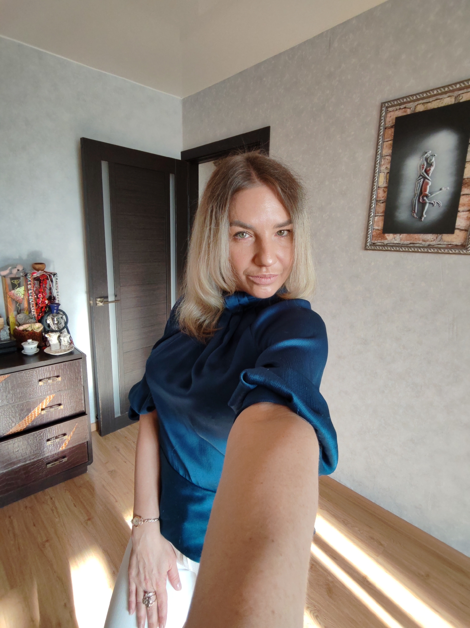 A woman with blonde hair and green eyes takes a selfie in what appears to be a residential room. She wears a dark blue blouse and white pants, looking directly at the camera with a calm expression. Her right arm is extended, holding the camera. She wears a gold-toned watch and rings on her left hand.
The room features light-colored, textured wallpaper and a light wood floor. Strong, angled sunlight streams onto the floor, indicating it is likely late morning or early afternoon. To the left, a dark brown dresser with a textured, crocodile-skin-like pattern and metallic handles holds various decorative items: a colorful hanging ornament, a small ornate blue and gold teapot or decorative object, and two white teacups with saucers. Behind the dresser, a dark brown door with a vertical frosted glass panel and a silver handle is partially visible. On the right wall, a framed artwork displays two intertwined figures, possibly dancers or lovers, on a dark background, set within a rustic, brick-patterned frame. No visible text is present in the image. The location context provided is Petropavlovsk-Kamchatsky, Russia.