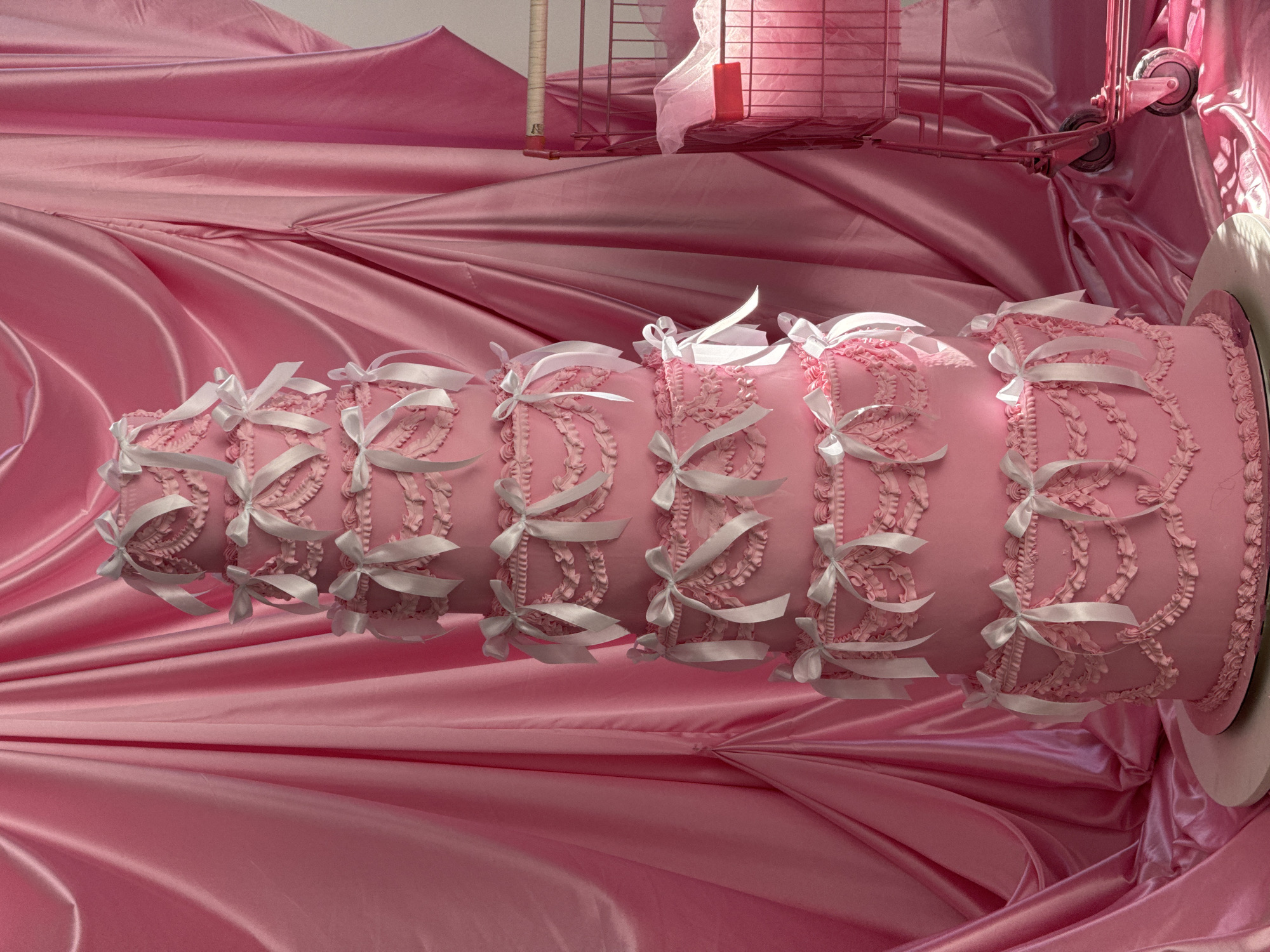 The image depicts a pink, multi-tiered cake decorated with white ribbons and frilled icing. It rests on a shiny, pink satin cloth. In the background, a pink wire shopping cart with wheels is visible. A shelf with a pink item sits above the cart. The location is Unknown, Unknown.