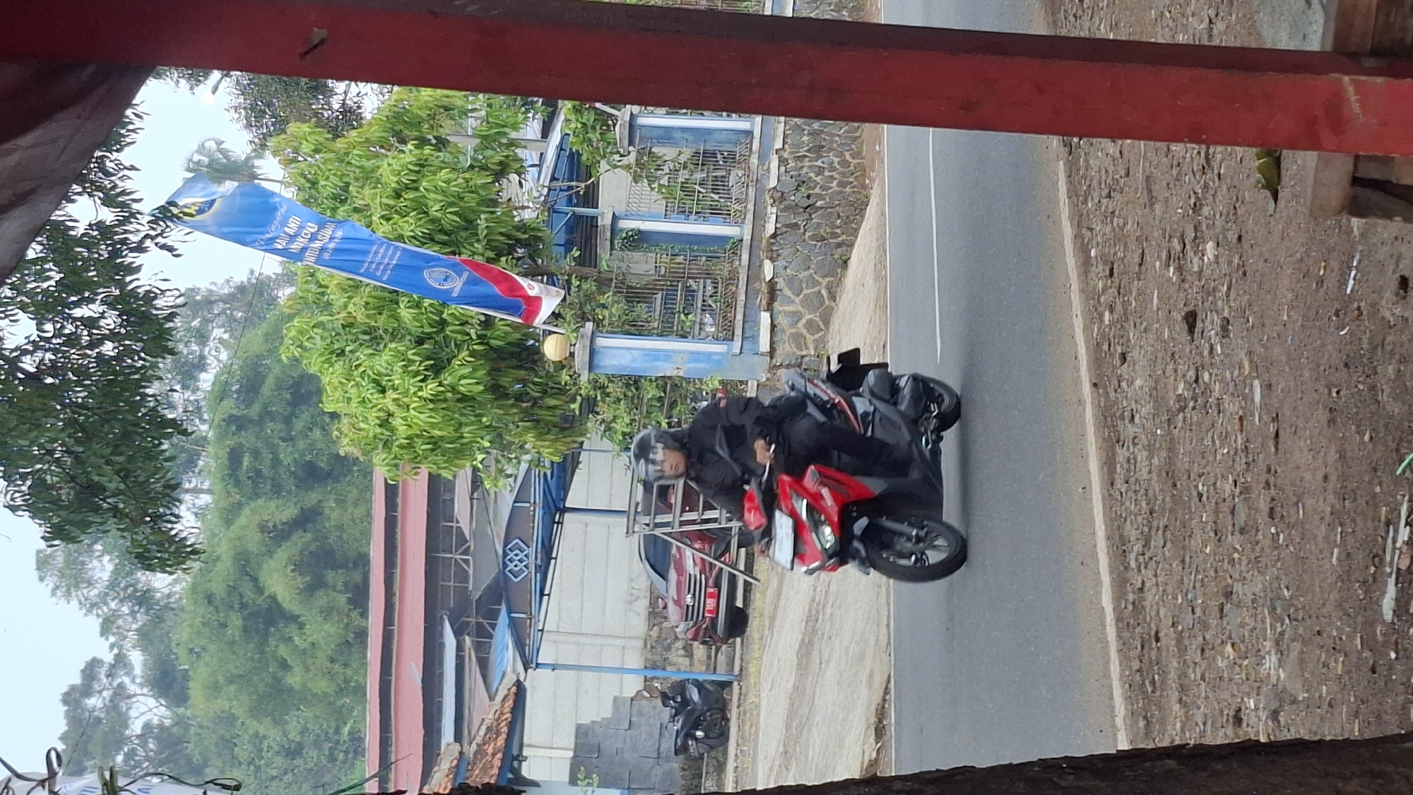 A motorcyclist wearing a black jacket and helmet rides a red and black motorcycle on a paved road. The motorcyclist is looking forward. In the background, there is a red car parked behind a white fence. A blue banner with white lettering hangs from a pole. Trees and buildings are visible in the distance. The scene appears to be in a semi-urban area, possibly in Cilaku, Indonesia, as indicated by the presence of tropical vegetation and the style of the buildings and banner. The road is asphalt with a white line marking the edge. The ground on the side of the road is covered with dirt, gravel, and some debris.