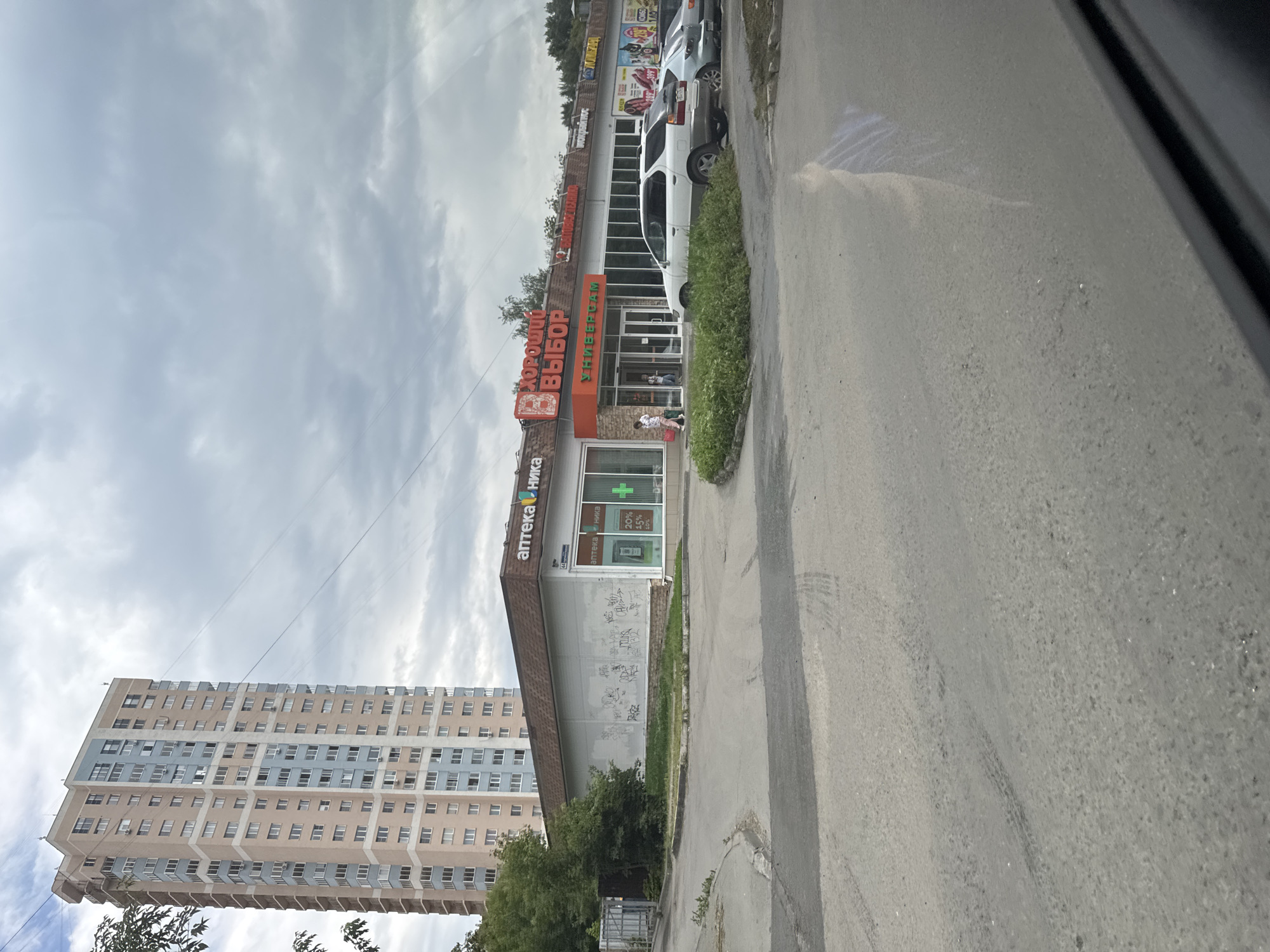 A high-rise apartment building stands tall in the background, its facade a blend of pinkish-beige and light blue. In the foreground, a low-rise commercial building houses various shops. One prominent shop, labeled "Хороший Выбор," features a bright orange sign with red lettering above its entrance. To its left, an "Аптека Ника" (Pharmacy Nika) sign is visible, with a green cross symbol and a discount percentage display. The shops are set back from a paved area, where several cars are parked, including a white sedan and a silver sedan. A patch of green bushes is located in front of the shops. The sky above is overcast with grey clouds. A road with tire marks is in the immediate foreground.