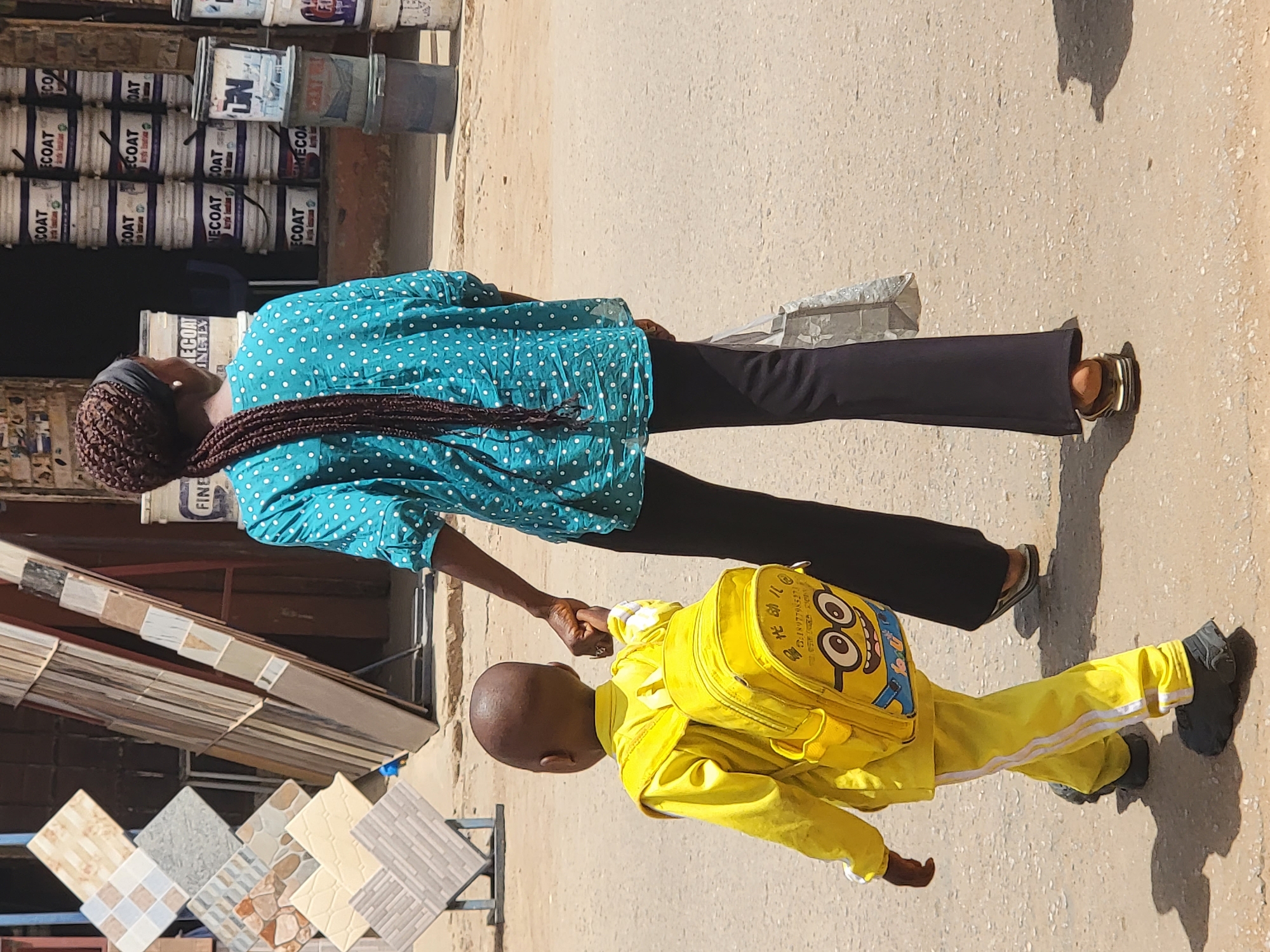 A medium shot shows a woman and a child walking on a street. The woman, seen from above, is wearing a turquoise shirt with white dots, dark pants, and sandals. Her hair is braided. The child, wearing a yellow tracksuit and a yellow backpack with a Minion design, holds her hand. The setting appears to be a commercial area in Maiburiji, Nigeria, with paint buckets and tile displays visible in the background, indicating the location may be near a store selling home improvement supplies.