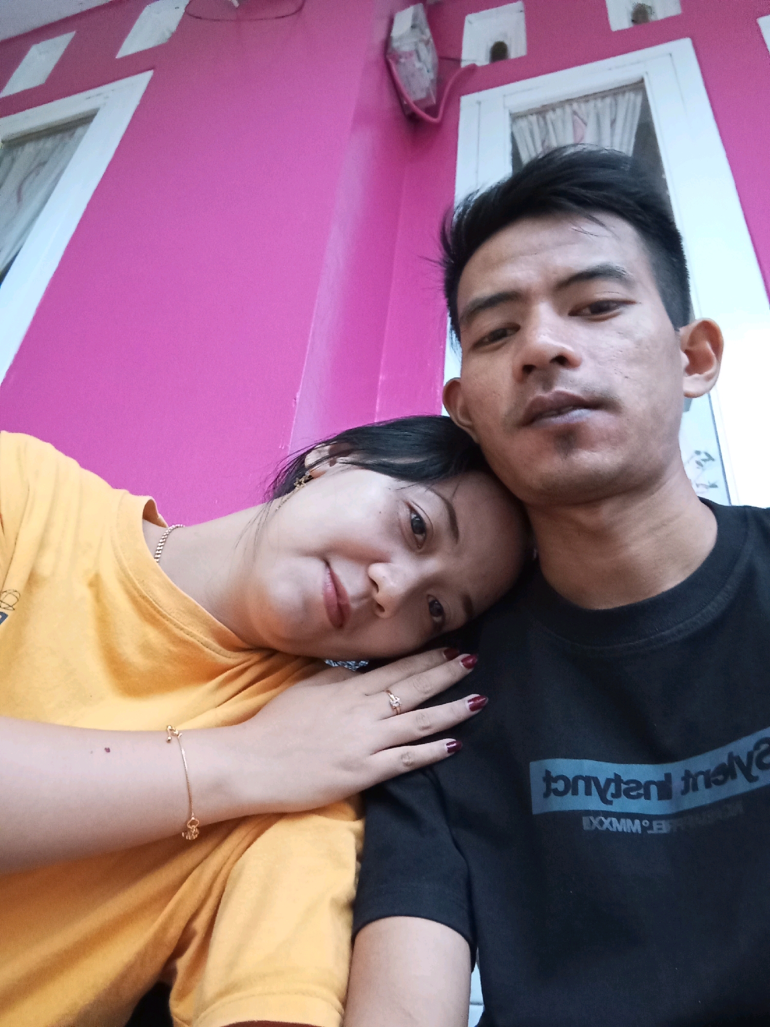 A close-up selfie shows a young man and a woman in front of a bright pink wall with white window frames and trim. The man is on the right, facing the camera with his eyes open and mouth slightly parted. He has dark hair and is wearing a black t-shirt with a graphic logo. The woman is on the left, resting her head on the man's shoulder and looking towards the camera with a slight smile. She has dark hair pulled back and is wearing a yellow t-shirt. Her left arm is visible, adorned with a thin gold bracelet and a ring on her index finger. The background wall is a vibrant pink, with architectural details suggesting an outdoor setting. The overall image captures an intimate moment between the couple.