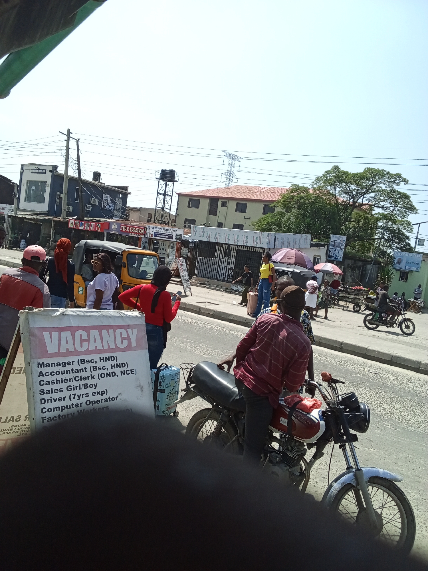 This is a street scene in Ado, Nigeria, depicting a busy thoroughfare with several individuals and vehicles. In the foreground, a man wearing a red and white striped shirt is seated on a motorcycle. To his left, a large white sign displays "VACANCY" prominently, followed by a list of job titles: Manager (Bsc, HND), Accountant (Bsc, HND), Cashier/Clerk (OND, NCE), Sales Girl/Boy, Driver (7yrs exp), Computer Operator, and Factory Workers.
Behind the sign, other people are visible. A person in a red shirt stands next to a yellow auto-rickshaw (keke napep). Other individuals are seen walking or standing on the sidewalk. Buildings line the street, with various shops and signs, including one that reads "JEP.K CREATION." Power lines crisscross the sky, and a water tower is visible in the distance. A red umbrella is set up near the auto-rickshaw. The overall environment suggests an active urban setting with commercial activity.