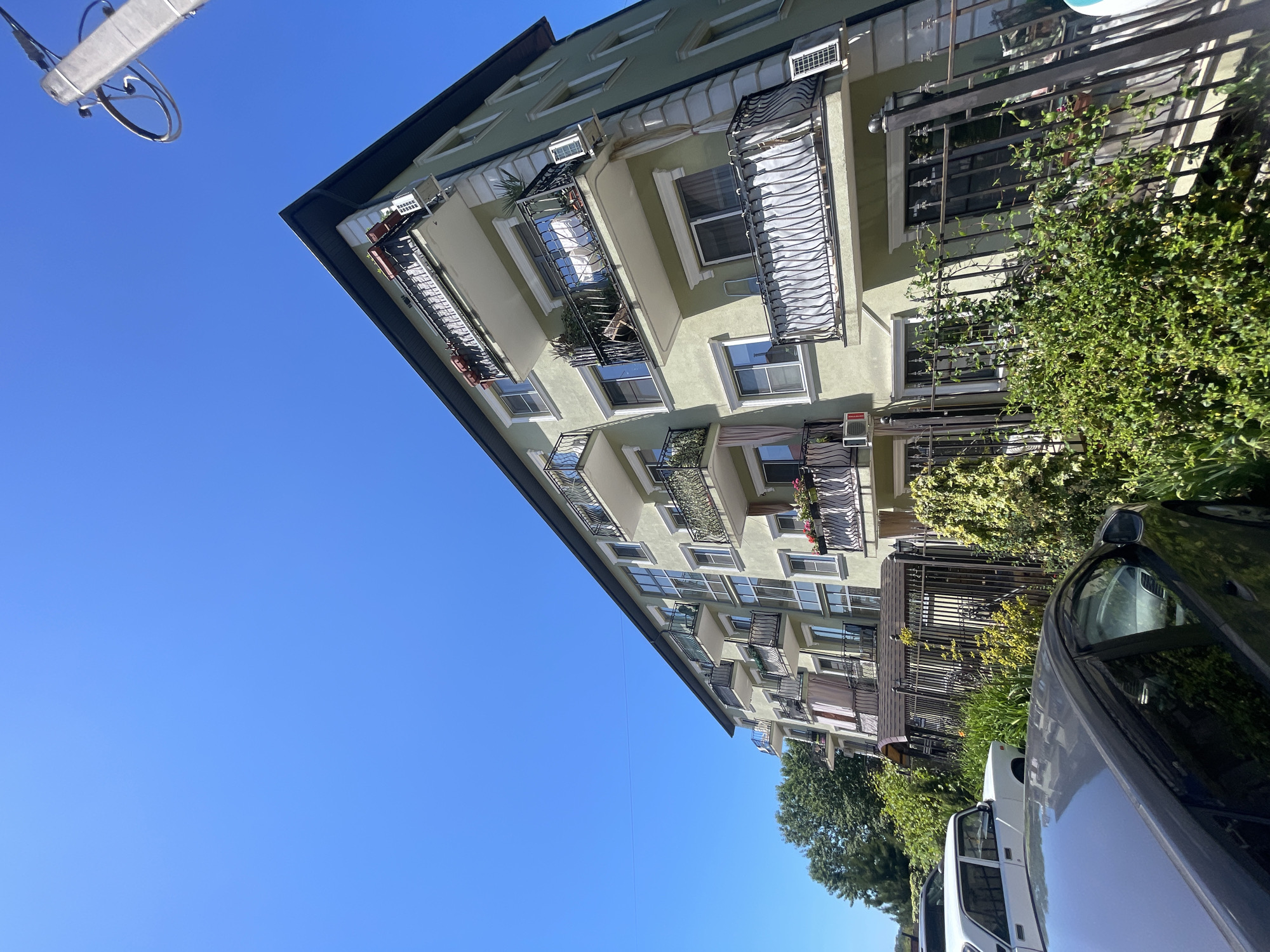 A tilted daytime shot captures a residential building with a bright, clear blue sky dominating the upper left portion of the frame. The multi-story building, painted in a light olive green or pale yellow hue, features numerous balconies adorned with ornate wrought iron railings. Several balconies have white awnings, and some hold potted plants or flower boxes, suggesting residential use. Air conditioning units are visible beneath several windows.
The building's architecture includes multiple windows, some with white frames, and a dark-colored sloped roofline visible at the top edge. Lush green bushes and trees line the foreground and parts of the right side, partially obscuring the lower levels of the building and a dark-colored sedan parked on the street. A white utility van or similar vehicle is partially visible behind the sedan. The foliage suggests a warm climate.
In the extreme top left corner, a piece of a light-colored utility pole or street light extends into the blue sky. The sunlight is bright, indicating either morning or afternoon, and there are no clouds in the sky. The overall scene depicts a typical street view in a warm, sunny location like Sochi, Russia, with the image intentionally rotated, giving a dynamic, off-kilter perspective to the otherwise static urban landscape. No visible text is discernable.
