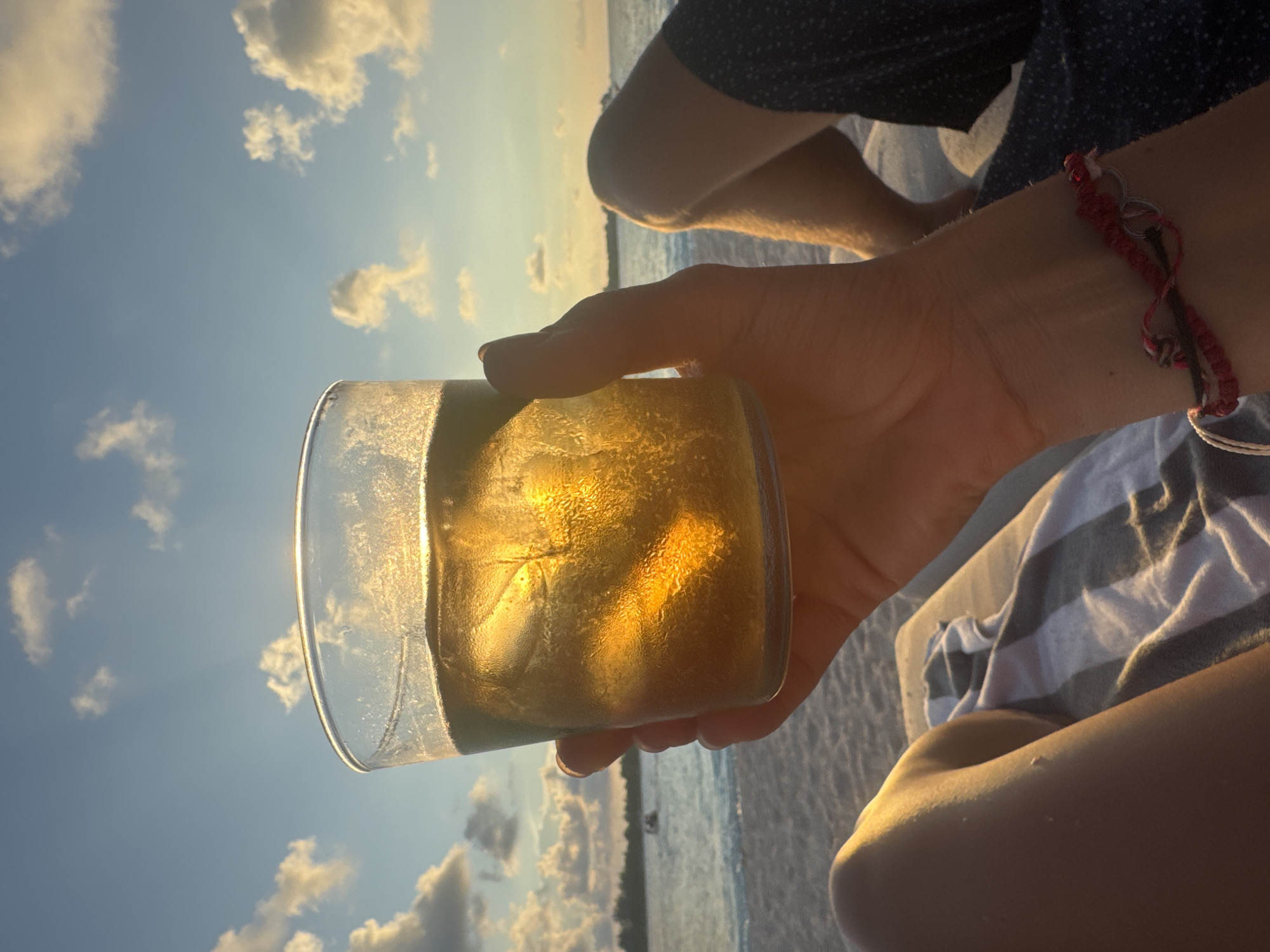 A person is holding a glass of amber-colored liquid, possibly a cocktail, against a backdrop of a beach and a cloudy sky at sunset. The beach appears to be in an unknown location in Indonesia, based on the sand and water. The person is wearing a red and white string bracelet on their wrist. The scene is lit by warm sunlight, creating a golden glow on the liquid in the glass and highlighting the clouds. The background includes another person's legs and a striped towel or blanket, suggesting a relaxed beach setting.