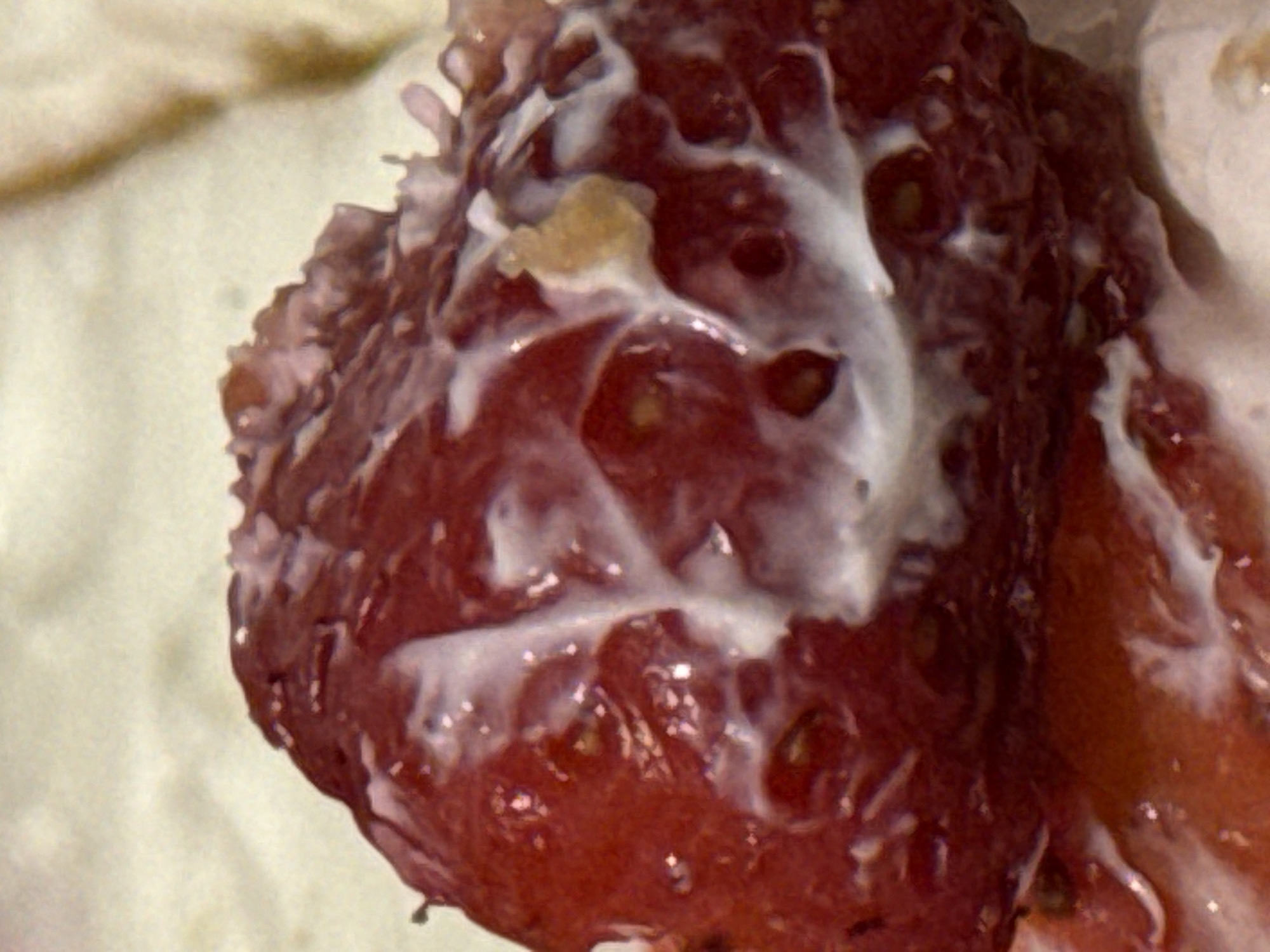 The image displays a close-up view of a strawberry that has been partially submerged in a white, creamy substance. The strawberry is deep red and shows visible seeds. The white substance appears to be liquid or semi-liquid, with some areas of thicker consistency, creating streaks and patterns across the surface of the fruit. A small, pale yellow object, possibly a piece of food debris or a dislodged seed, is visible on the strawberry. The background is out of focus and appears to be a light-colored surface. There are no indications of a specific location in Rustavi, Georgia.