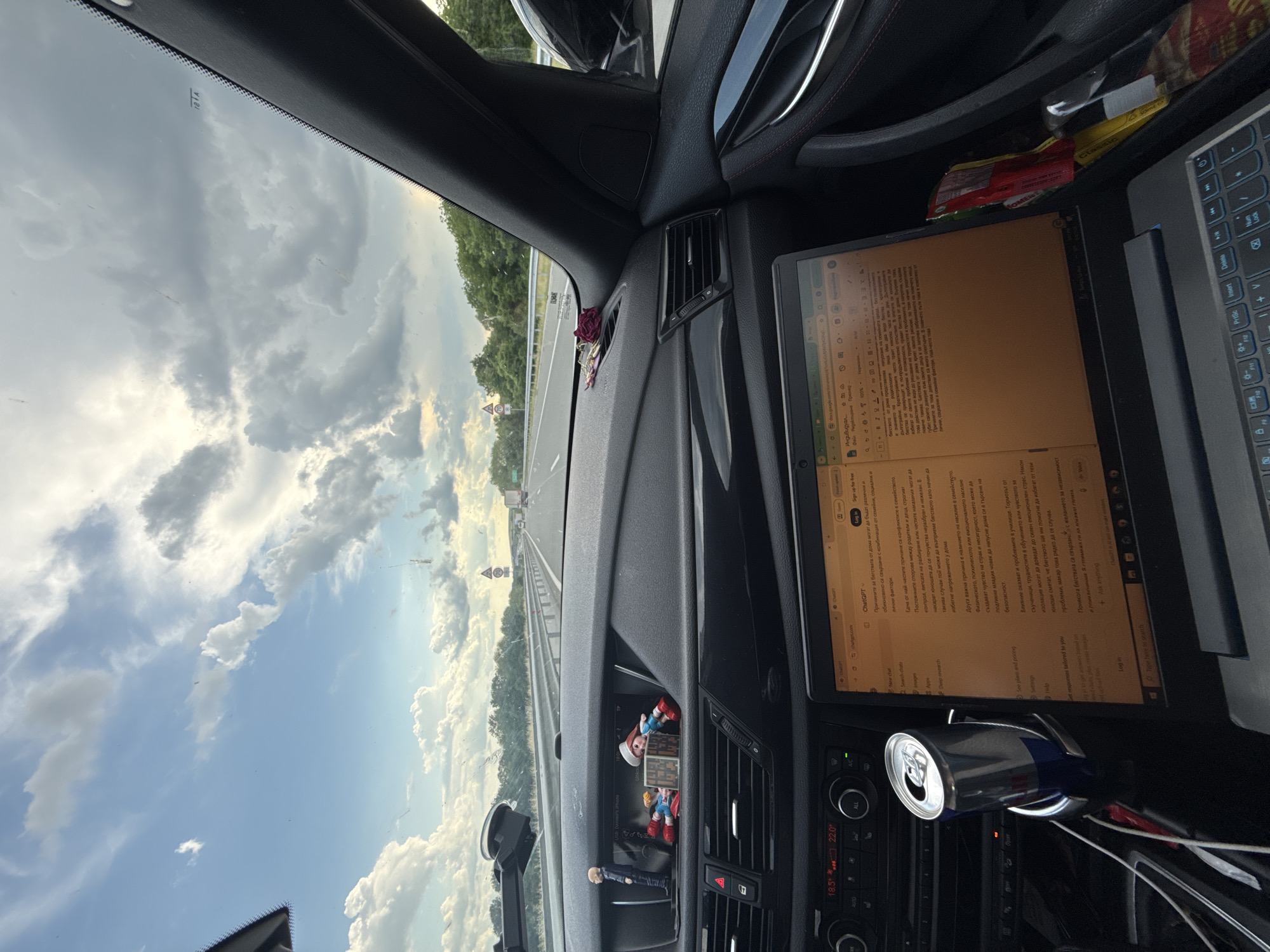 The image shows the interior of a car driving on a highway. The dashboard is visible, with a laptop open and displaying text, a can of energy drink in a cup holder, and several figurines on the dashboard. The exterior view through the windshield shows a highway with trees and cloudy sky. The location is identified as Arnautito, Bulgaria. There are no people visible in the image. The scene depicts someone working on a laptop while traveling in a car.