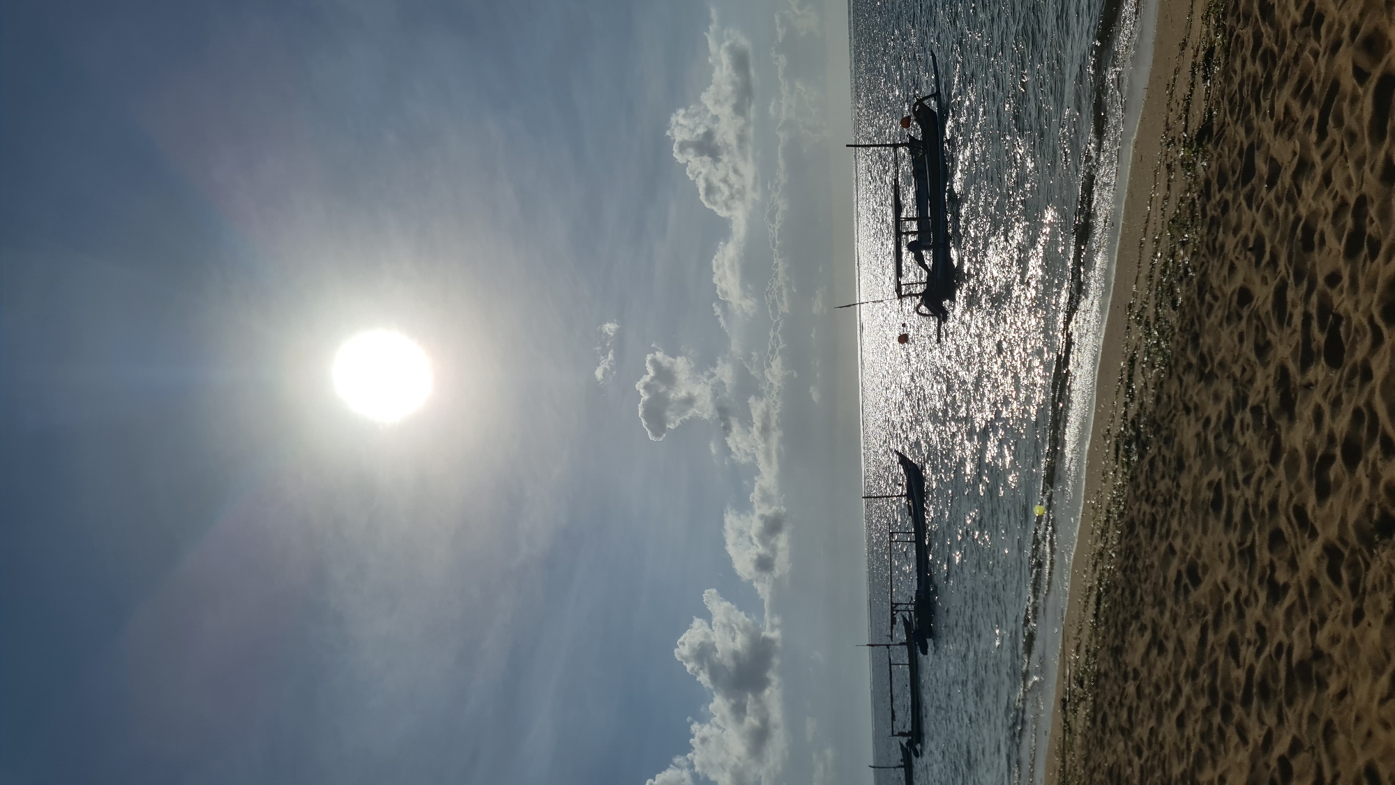 This is a vertical, medium-shot photograph taken at Pantai Sanur, Denpasar, Indonesia. The sun is high in the sky, partially obscured by thin clouds, casting a bright glare on the water. Several traditional Balinese fishing boats, with their characteristic outriggers and thatched roofs, are moored in the calm, shallow ocean. The foreground is dominated by a sandy beach, with some seaweed and debris washed ashore. Gentle waves lap the shoreline. The overall scene depicts a tranquil morning or afternoon at a popular tourist beach.