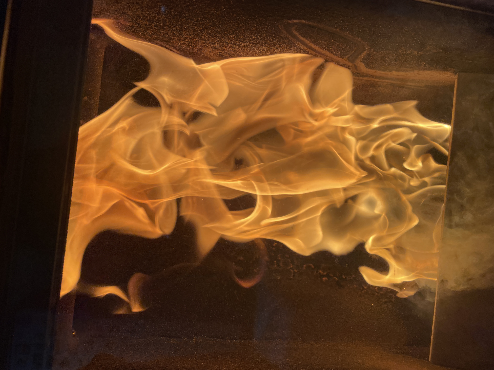 The image shows a fire burning inside a fireplace or stove. The flames are a bright orange-yellow color and fill the majority of the frame. The interior of the fireplace is dark. The location is unknown.