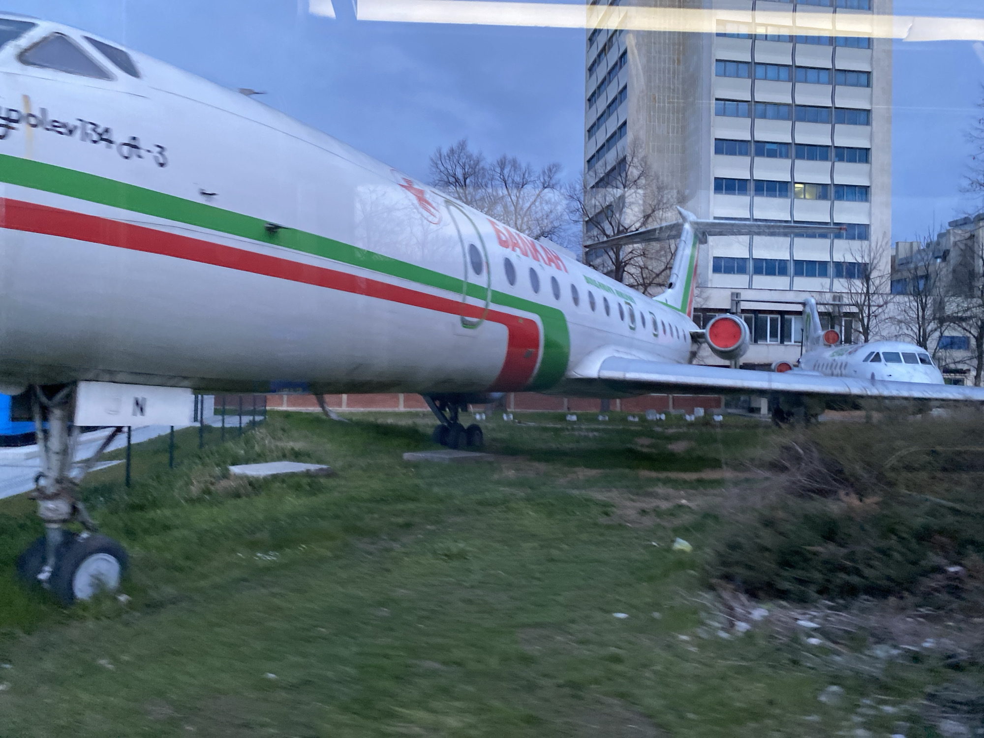 The image shows two Tupolev Tu-134 airplanes on display in a grassy area. The planes are painted white with red and green stripes, and the word "Балкан" (Balkan) is visible on the side of one of the planes. The plane in the foreground has the text "Tupolev 134 A-3" printed on the nose. In the background is a tall, modern building with many windows and bare trees. The scene appears to be captured from behind a window, causing reflections.
The location is likely somewhere in Sofia, Bulgaria, based on the airplane's Balkan Bulgarian Airlines livery, a former Bulgarian state airline.