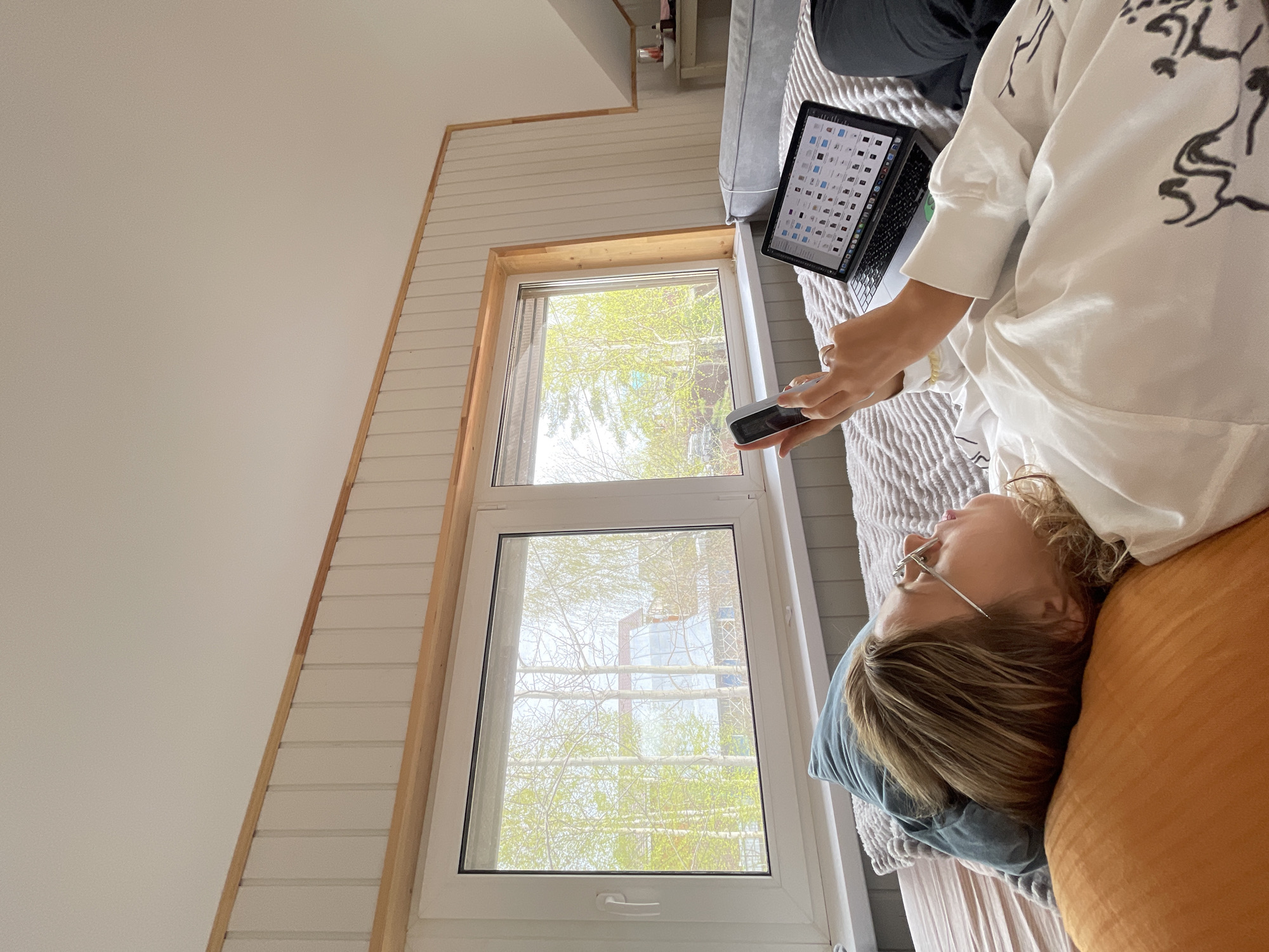 A young woman wearing glasses and a white shirt with black designs is lying on a bed. She is using a small handheld device in her right hand, and a laptop computer rests next to her on the bed, displaying a screen filled with icons. Pillows are positioned behind her head. The setting appears to be indoors, with a window overlooking trees and what seems to be a construction site in the distance. Based on the visual cues of the architecture and landscape, it is likely that the location is Sestroretsk, Russia.