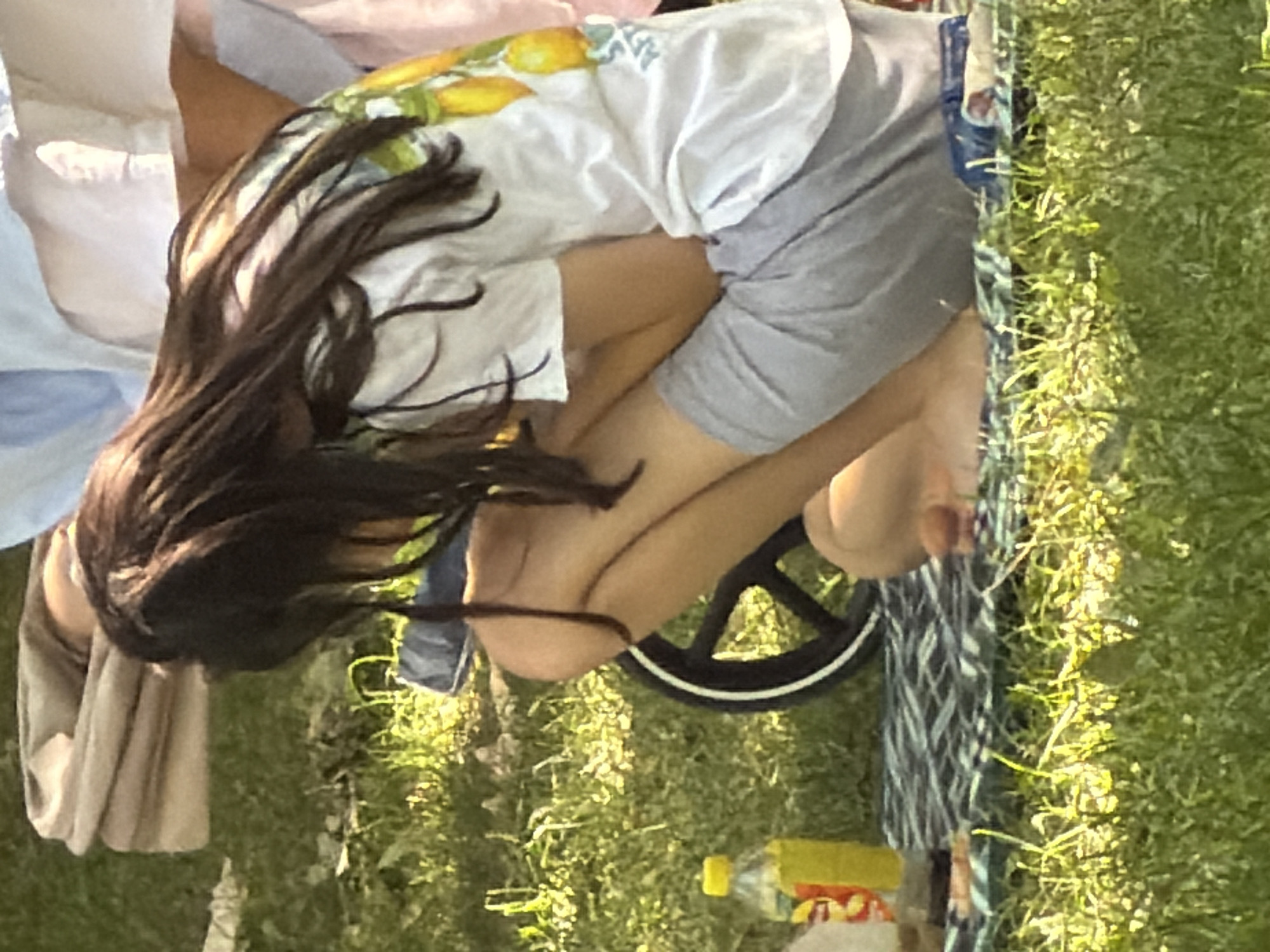 A young girl with long dark hair is sitting on a checkered blanket on grass. She is wearing a white t-shirt with a lemon print and grey shorts. Her legs are crossed, and her bare feet are visible. A bottle of yellow liquid with a yellow cap is next to her on the blanket. In the background, there is a folded blanket or fabric and a black wheel with a white stripe, possibly part of a stroller or cart. The scene appears to be an outdoor setting in a park or yard, with sunlight filtering through the grass. The overall impression is one of a casual outdoor activity.