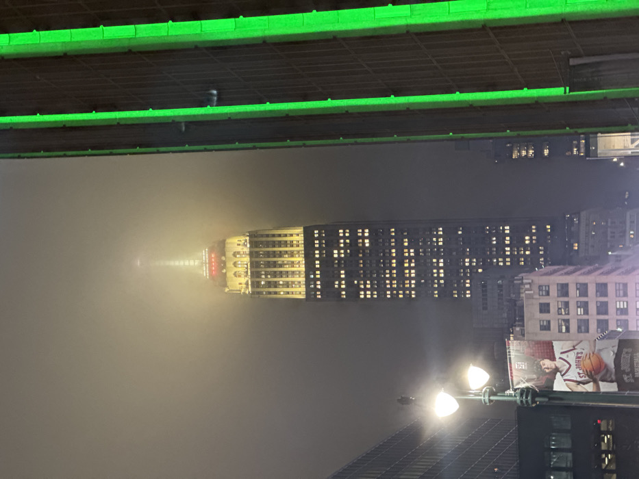 The Empire State Building rises through a dense fog, its top illuminated. The building's windows are lit, creating a grid-like pattern. A green light structure is visible above, possibly a bridge. Streetlights illuminate the foreground. An advertisement featuring a basketball player is on a nearby building. The fog obscures the surrounding cityscape, giving the scene an ethereal quality.