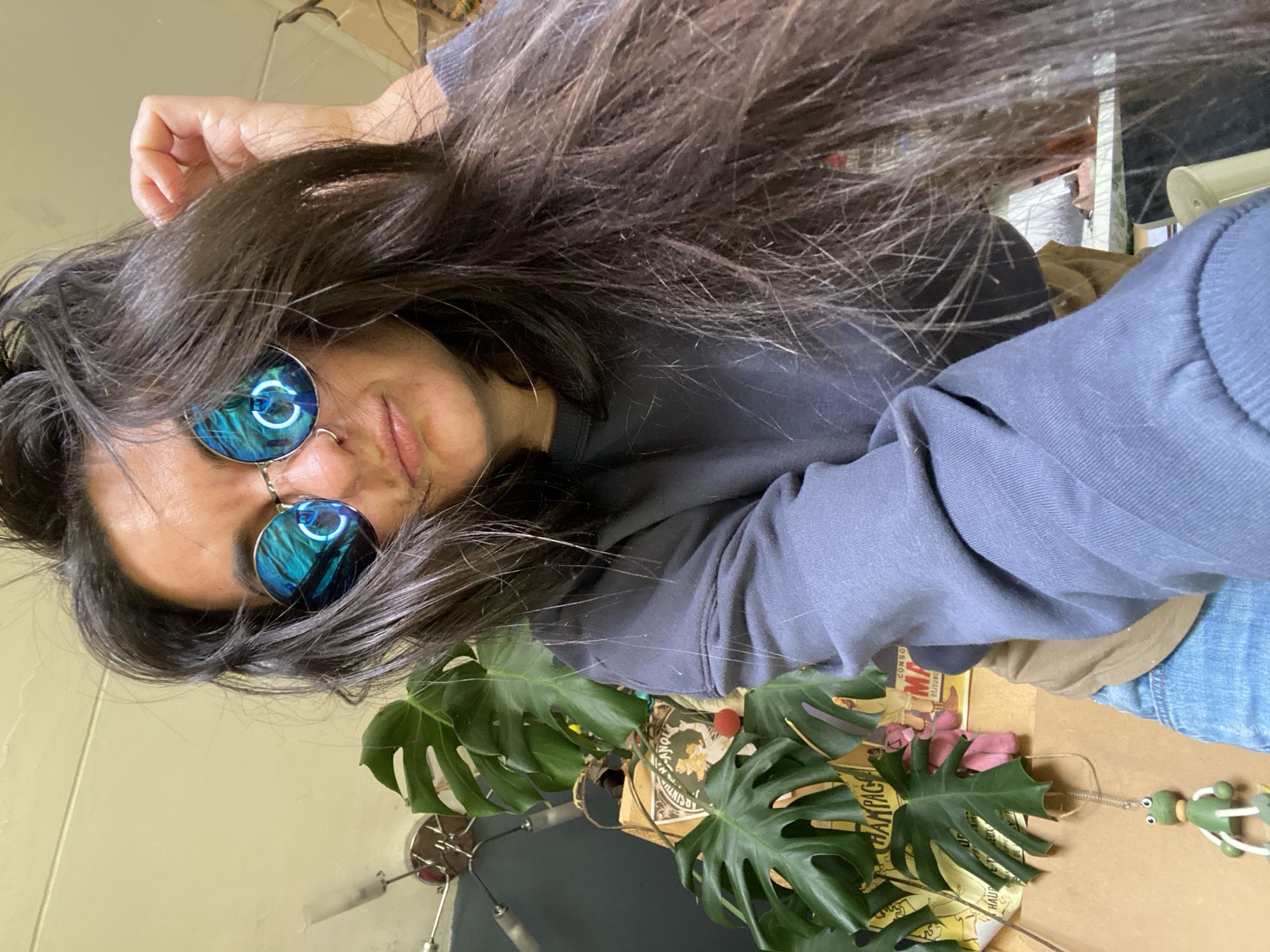 A woman with long brown hair and blue mirrored sunglasses is the primary subject. She is wearing a dark blue sweatshirt. Her face is partially obscured by her hair. In the lower part of the image, there's a Monstera plant, a wooden frog toy, and a stack of papers. The location is not discernible and cannot be identified as Moscow, Russia.