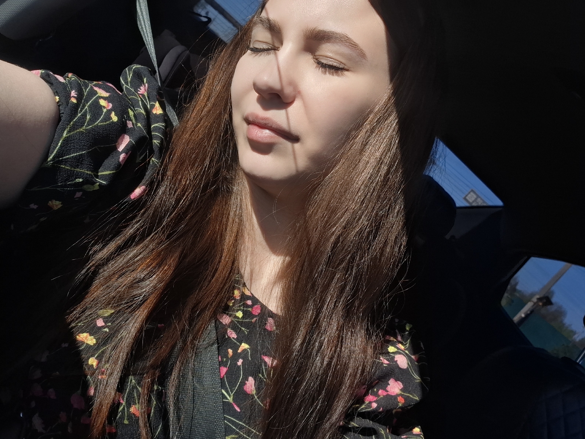 A woman is in a car. She has long brown hair, closed eyes and fair skin. She is wearing a black dress with small pink and yellow flower patterns and a seatbelt. The car is likely in Chaikovsky, Russia, given the image was sourced from there. The scene is in sunlight.