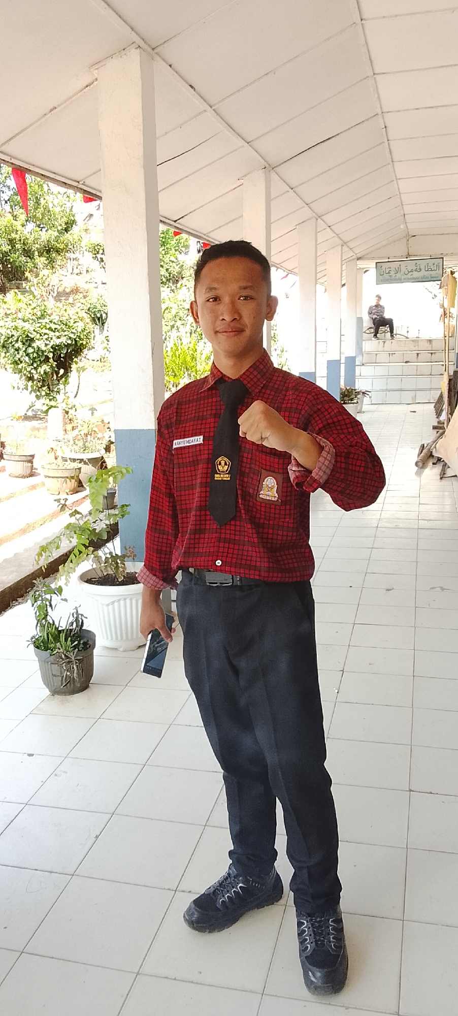 A young male stands in a covered walkway, holding a smartphone in his left hand and clenching his right fist. He wears a red and black plaid long-sleeved shirt, a black tie with a school emblem, and dark pants. His black sneakers are visible. The walkway has white tiled flooring and a white ceiling. Green plants in white and grey pots are situated to his left. In the background, another person is seated on steps. A sign with Arabic script is mounted on the wall. The location is identified as Jepara, Indonesia.