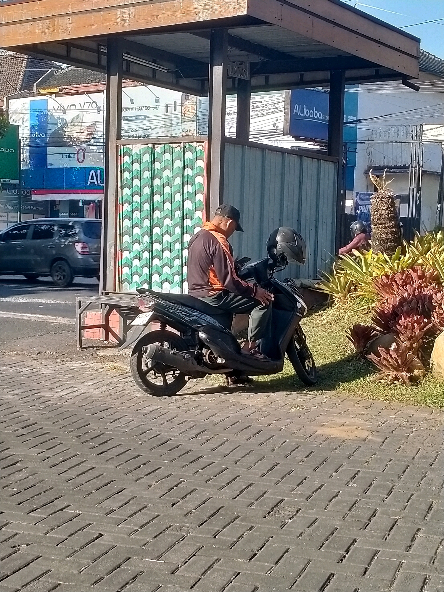 In the city of Batu, Indonesia, a man wearing a black baseball cap, an orange and black jacket, and dark pants is sitting on a black scooter. The scooter has a black helmet on the handlebars and is parked on a paved area with a distinctive interlocking brick pattern. Behind the man and scooter is a bus stop shelter with a wooden roof and metal supports. To the left of the shelter, there are advertisements for Vivo and Alibaba. A gray SUV is visible in the background, driving on a street. To the right of the shelter, there are lush green and red plants, a palm tree, and another person wearing a maroon shirt and helmet on a scooter. The scene appears to be a public space on a sunny day.