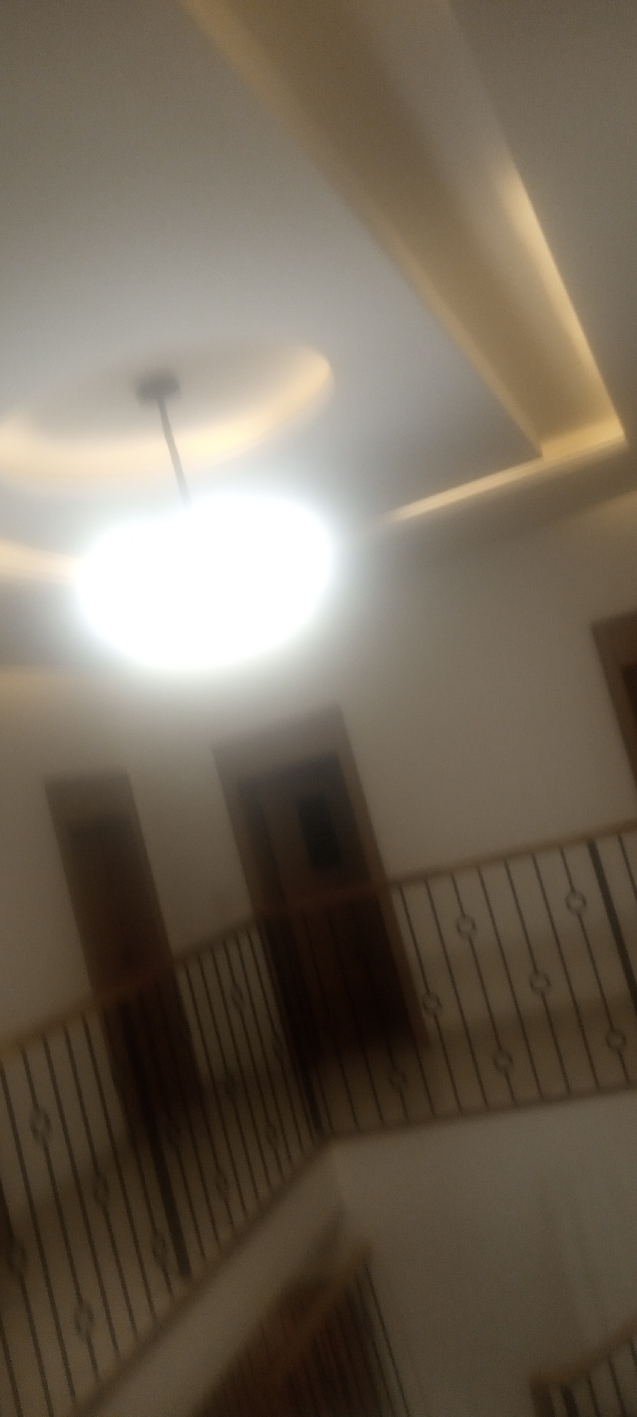 The image shows an interior space with a staircase and a railing with decorative metalwork. Two doors are visible on the upper level, flanking the railing. The ceiling features a circular light fixture and a decorative recessed design with indirect lighting. The overall color scheme is muted, with beige or cream walls and brown wood finishes. The scene appears to be in a private residence or similar building. The location in the city of Unknown, Unknown is unknown.