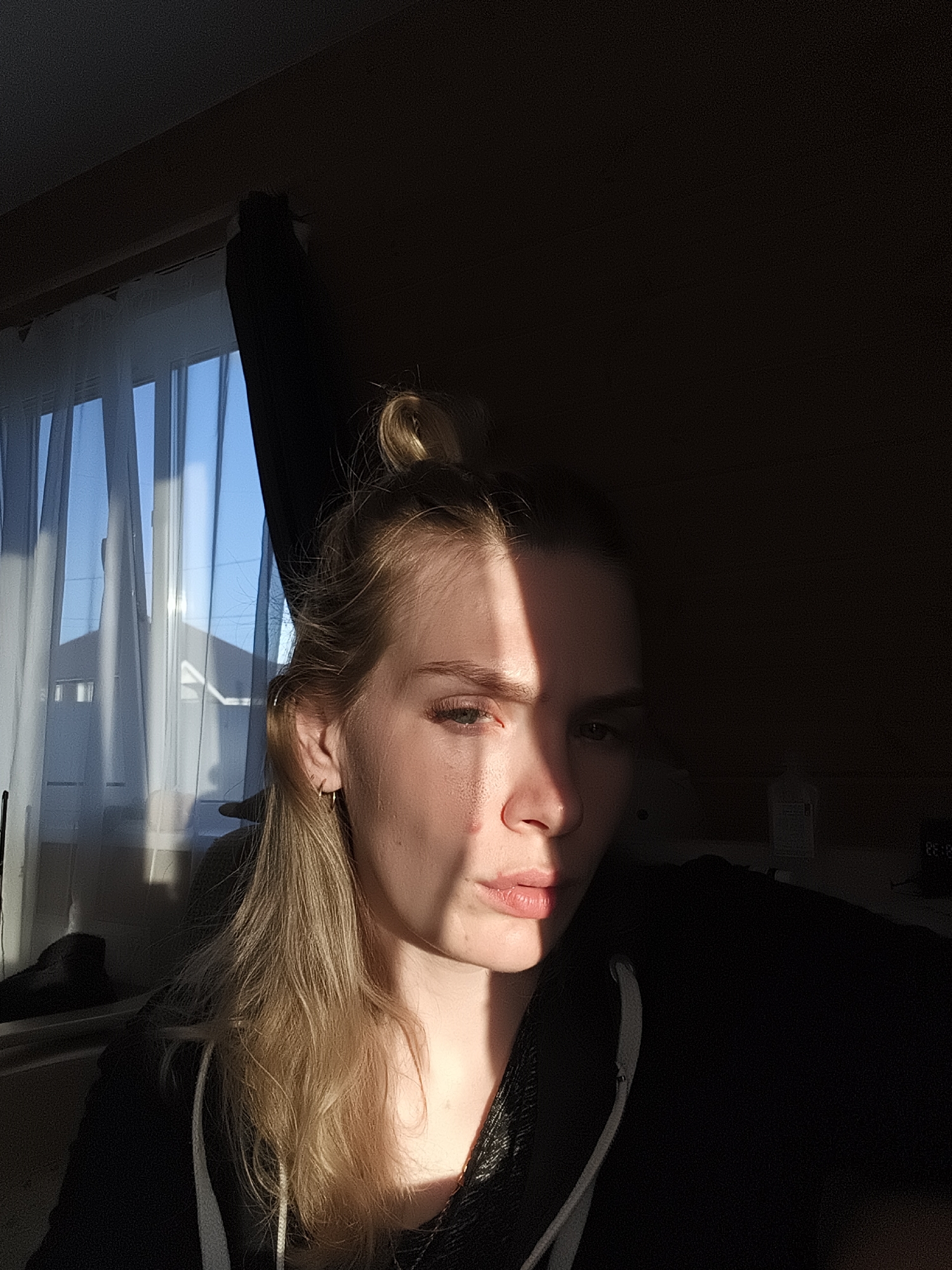 A photograph shows a person with blonde hair in a bun, wearing a black hoodie. Part of their face is brightly lit by sunlight, while the other part is in deep shadow. A window with white curtains is visible in the background, showing a view of rooftops. It is not possible to confirm the location in Русский Вожой, Russia, from the image.
