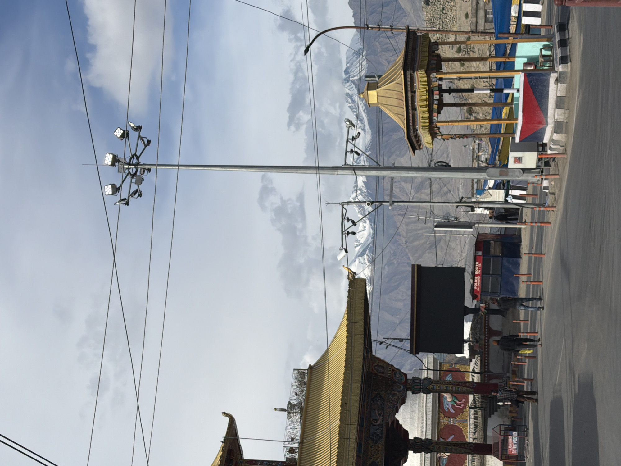 A wide shot shows a view of a street in Leh, India, with a mountainous backdrop. In the foreground, a road curves to the left. On the right side of the road, there is a small, open-air pavilion with a golden roof and colorful prayer flags. To the left of the pavilion is a modern street lamp with multiple floodlights.
Behind the pavilion, to the left, is a large structure with a golden, tiered roof, reminiscent of Tibetan architecture. This structure is supported by ornate pillars. In front of this building, a dark, rectangular screen is mounted. Further left, a white stupa-like structure is visible.
In the middle ground, a police booth with a red and blue facade is situated. Several people are walking on the street. One person in a plaid shirt and brown pants stands near a red and white barrier. Two other individuals are walking away from the camera.
The background is dominated by a range of imposing mountains with snow-capped peaks. The sky is partly cloudy. Electrical wires crisscross the frame, and several poles with lights and cameras are visible throughout the scene. The overall impression is one of a bustling, yet serene, mountain town.