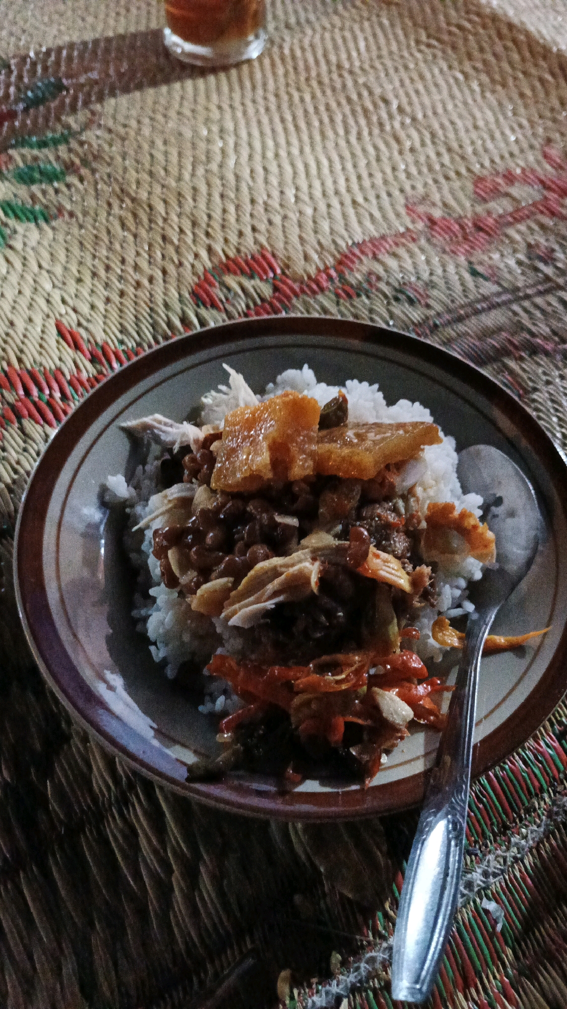 A plate of food, likely rice with various toppings, is centered on a woven mat. The plate contains white rice topped with shredded chicken, a reddish-orange stir-fry, and pieces of what appears to be fried pork skin or crackling. A spoon rests on the right side of the plate. In the upper background, a glass of dark liquid, possibly tea or coffee, is visible. The setting appears to be outdoors or in a rustic indoor environment, with the woven mat suggesting a traditional setting in Surakarta, Indonesia.