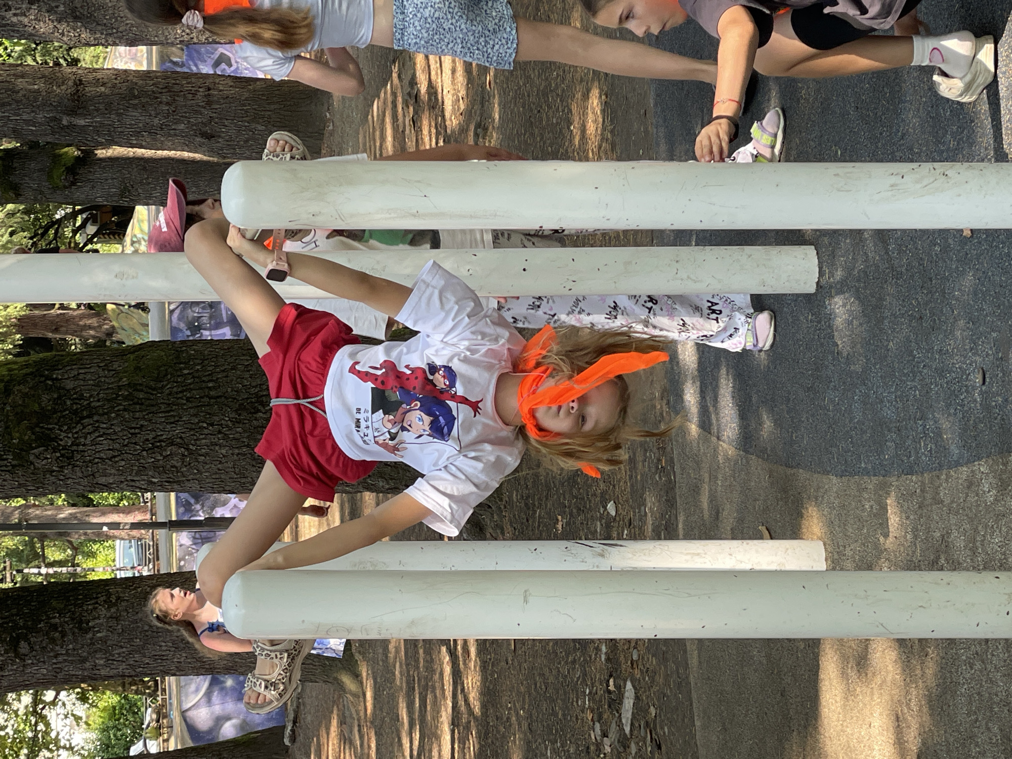 The image shows a group of children at an outdoor gym in Sochi, Russia. A young girl is hanging upside down from parallel bars, her legs spread wide. She is wearing a white t-shirt with cartoon characters, red shorts, and an orange bandana around her neck. Another girl in a blue dress stands in the background to the left, and a girl in a white t-shirt and red cap is visible on the right. Further to the right, a girl in a light blue patterned skirt and a white top stands. In the foreground on the right, another child is partially visible, wearing a dark t-shirt and black shorts. The gym appears to be set up in a park with trees and shadows visible on the ground.