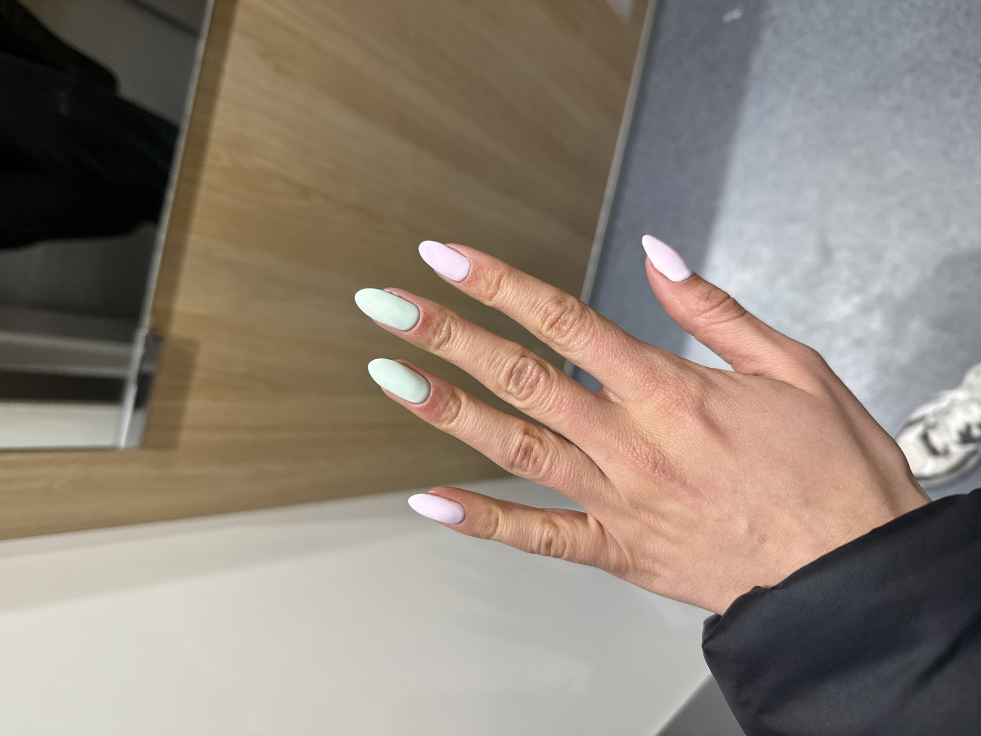 A right hand is presented against a background of furniture and floor. The hand has five fingers with almond-shaped nails, alternating between light green and light pink nail polish. The index and ring fingers are painted light green, while the thumb, middle, and little fingers are painted light pink. The hand is positioned in a slightly angled manner, fingers extended, with the wrist partially obscured by the sleeve of a dark-colored garment. The backdrop includes a light brown wooden surface, white furniture, and a grey floor. Partial view of a white sneaker and reflective surface are also visible in the background. The image's location in Sofia, Bulgaria cannot be verified from the available visual information.