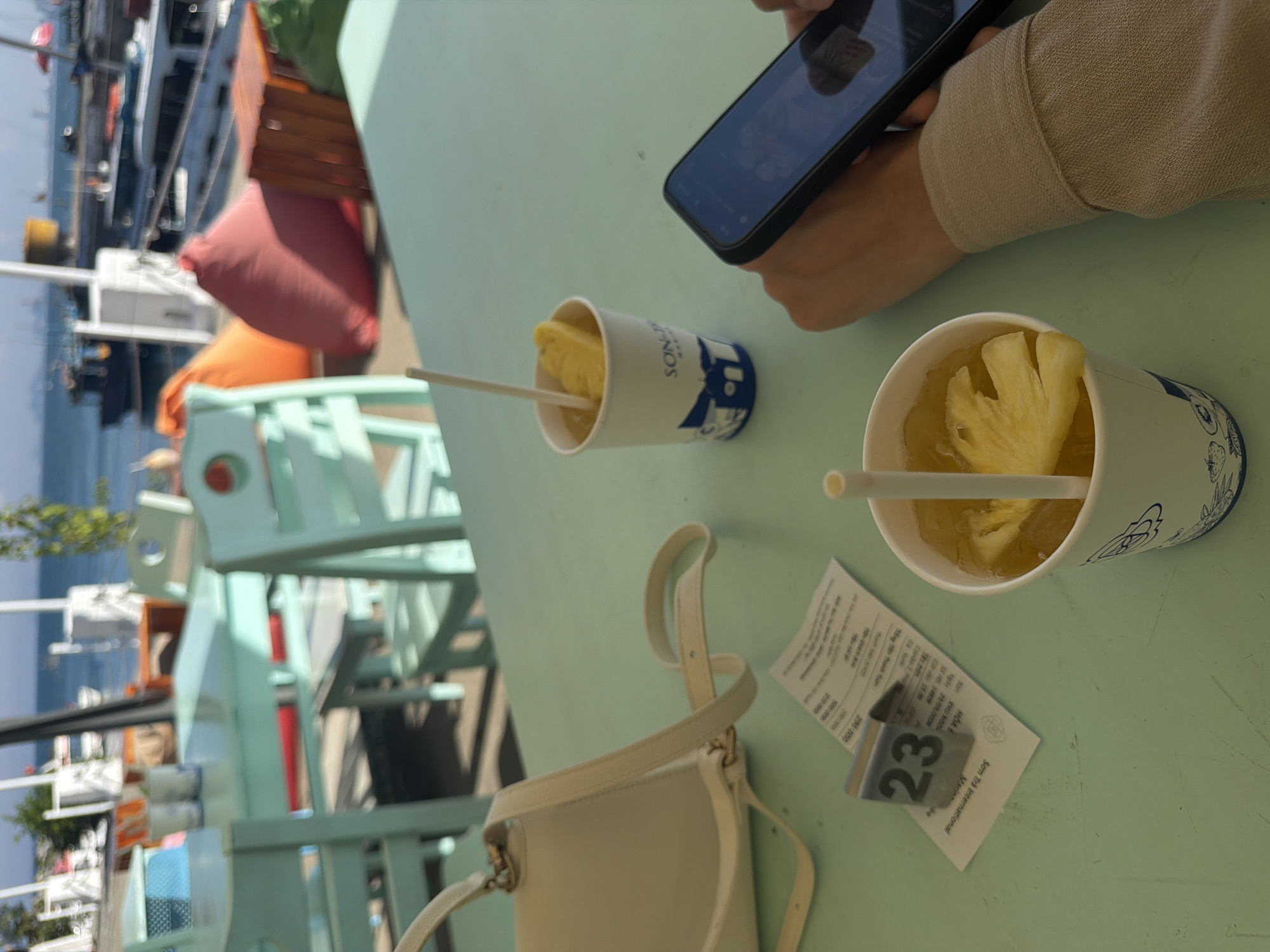 This image captures an outdoor setting, likely at a cafe or eatery in the city of Đà Nẵng, Vietnam. A light green table surface is the focal point, displaying a range of items. At the upper right corner, a person is holding a smartphone. Two cups of what appears to be pineapple juice or smoothie, each containing a straw, sit nearby. A small off-white purse with a thin strap is on the table's edge, along with a paper receipt and a table number stand reading "23".
The background reveals a blurry view of the city's waterfront, with boats and buildings in the distance. A turquoise chair with red accents is also visible. The overall impression is a casual, sunny day scene at a dining establishment in Đà Nẵng. The text on the receipt indicates "Son Trå International", which may be the name of the establishment.